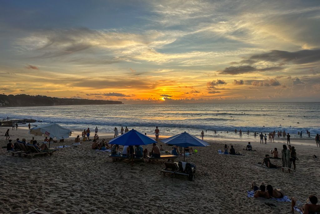 Sunset over a crowded beach. People relax under umbrellas and by the water, with waves in the background.