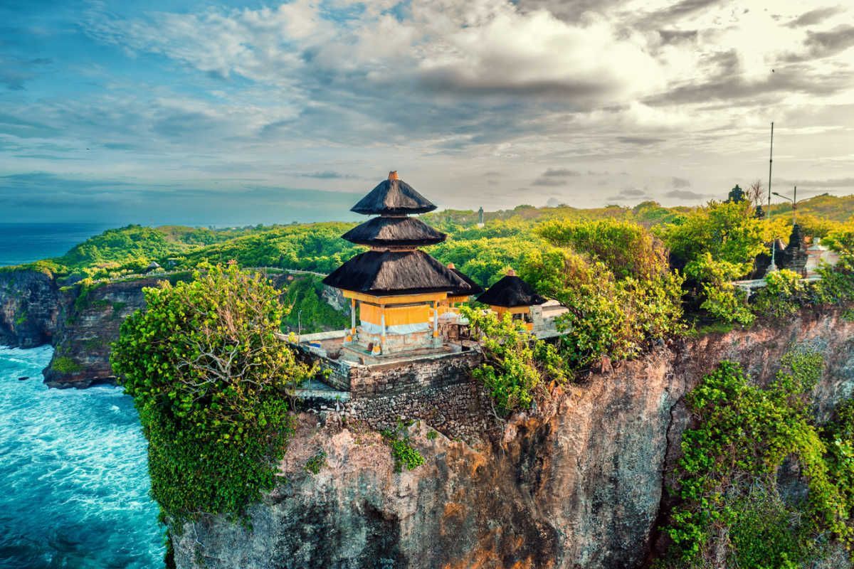 Temple perched on a cliff overlooking the ocean in Bali, Indonesia. Lush greenery, blue water, and cloudy sky.