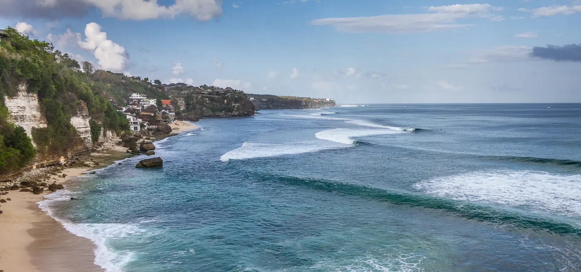A coastal view of a beach with cliffs and buildings, clear blue ocean and waves rolling towards the shore.