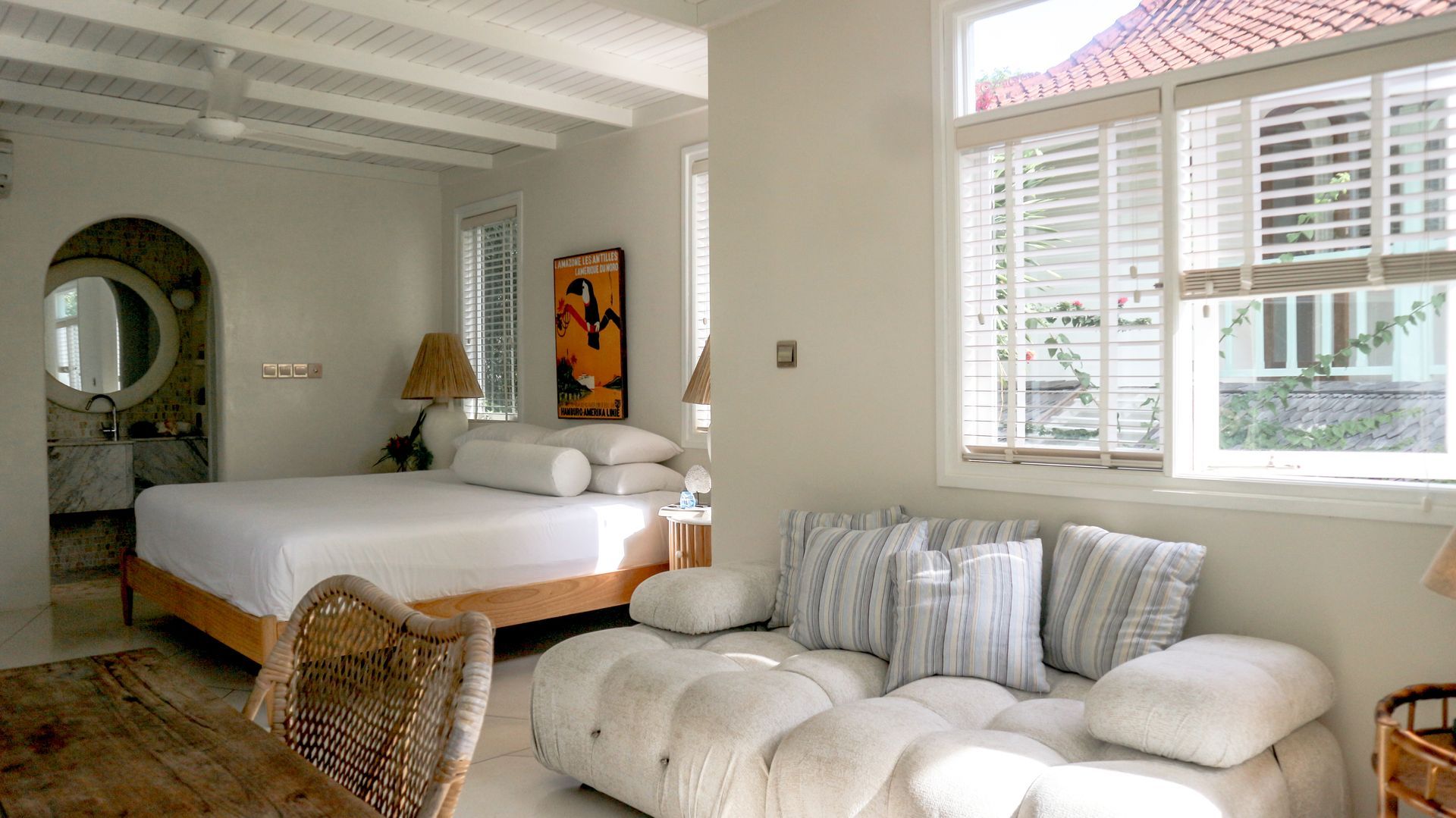 Bright bedroom interior with a white tufted couch, bed, and partially shuttered window; a bathroom doorway is visible.