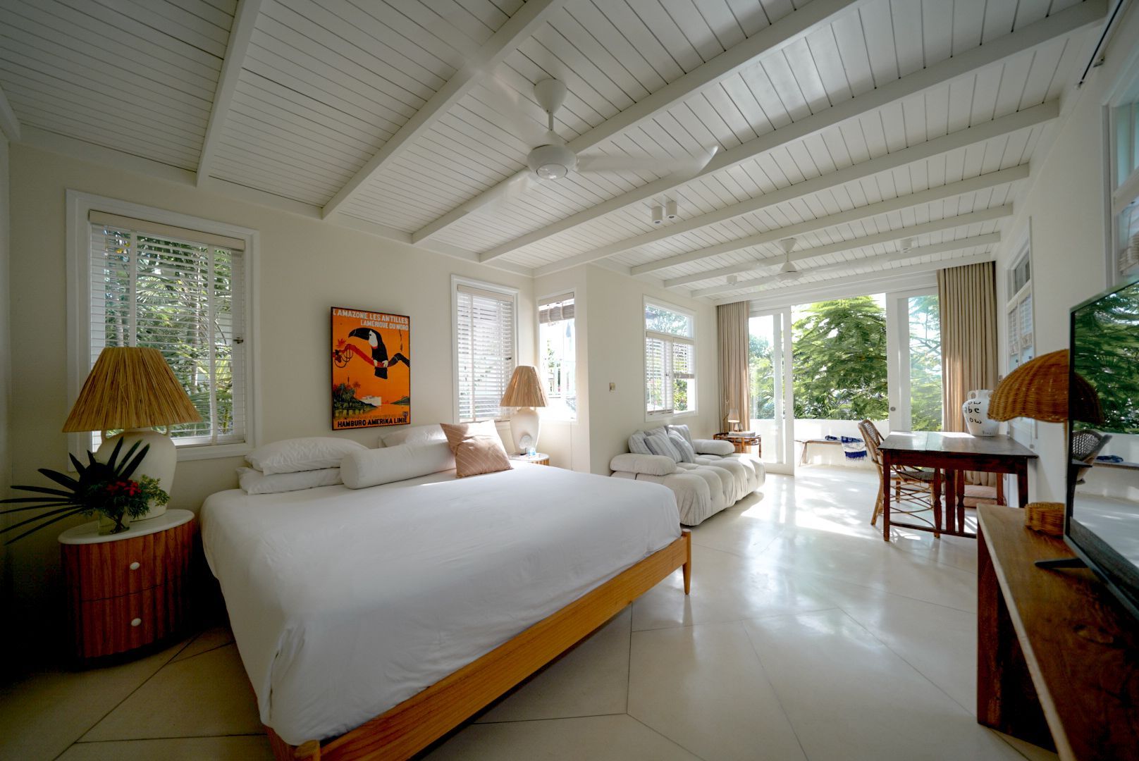 Bright, airy bedroom with white walls and wood ceiling. Features a bed, couch, and desk, with doors opening onto a patio.