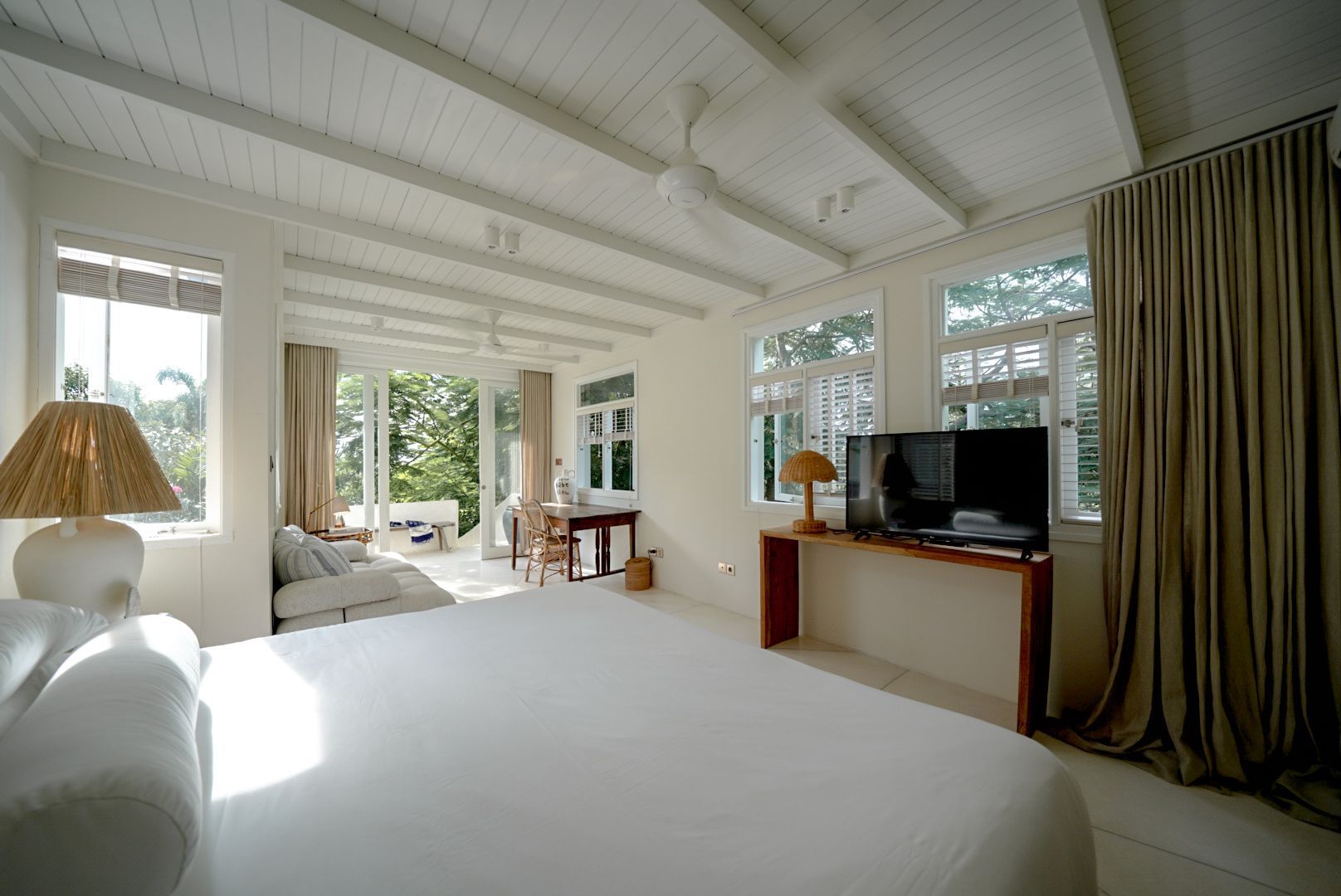 Bright bedroom with a white bed, ceiling beams, windows, and a view of greenery. A TV sits on a wooden console.