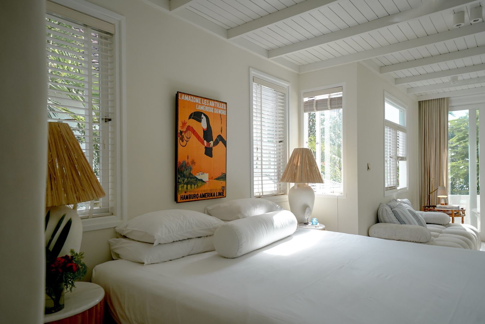 Bright bedroom with white bedding, windows, and a toucan poster. Sunlight streams in, illuminating the space.