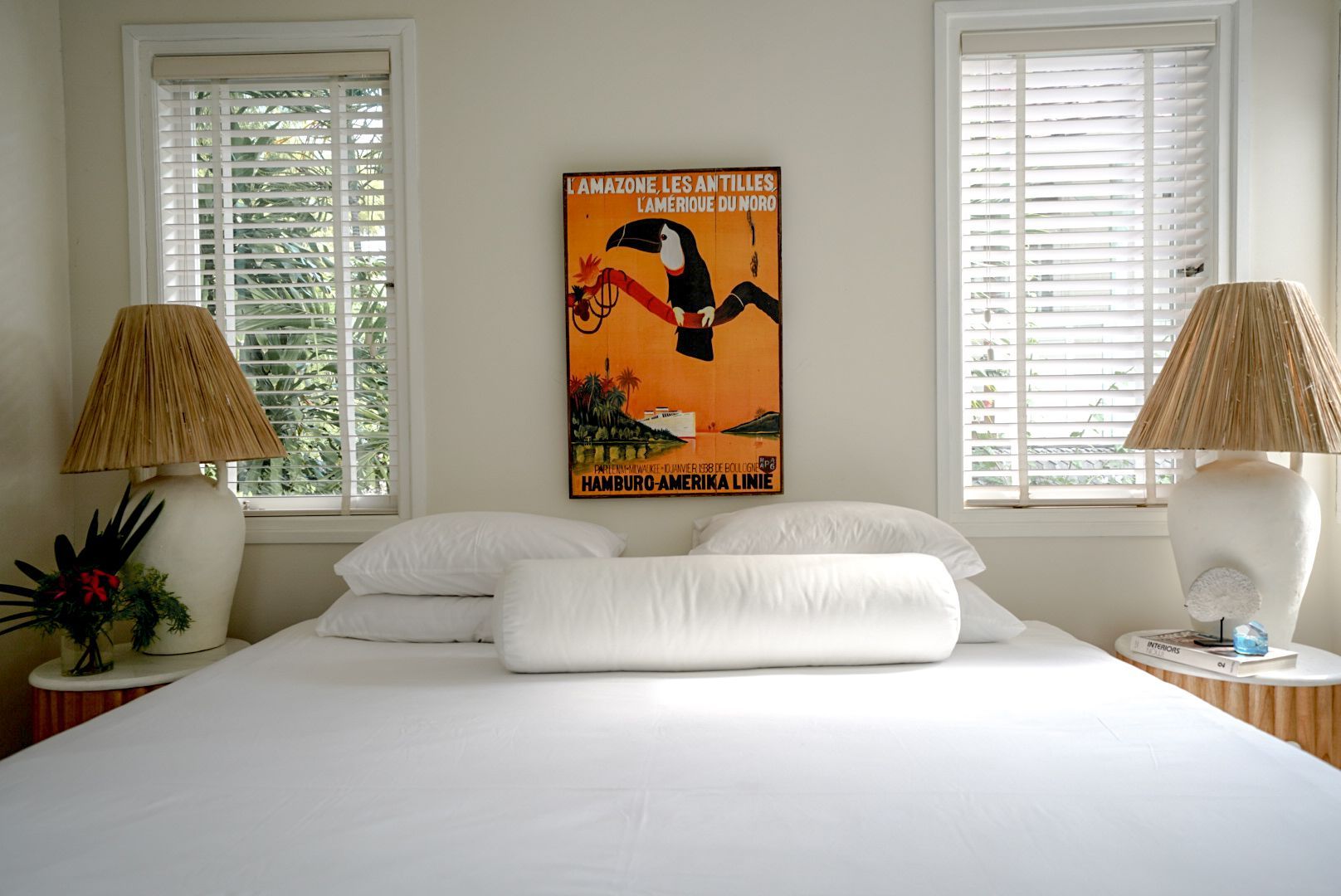 Bedroom with white bed, orange toucan poster, two windows with blinds, and lamps on either side.