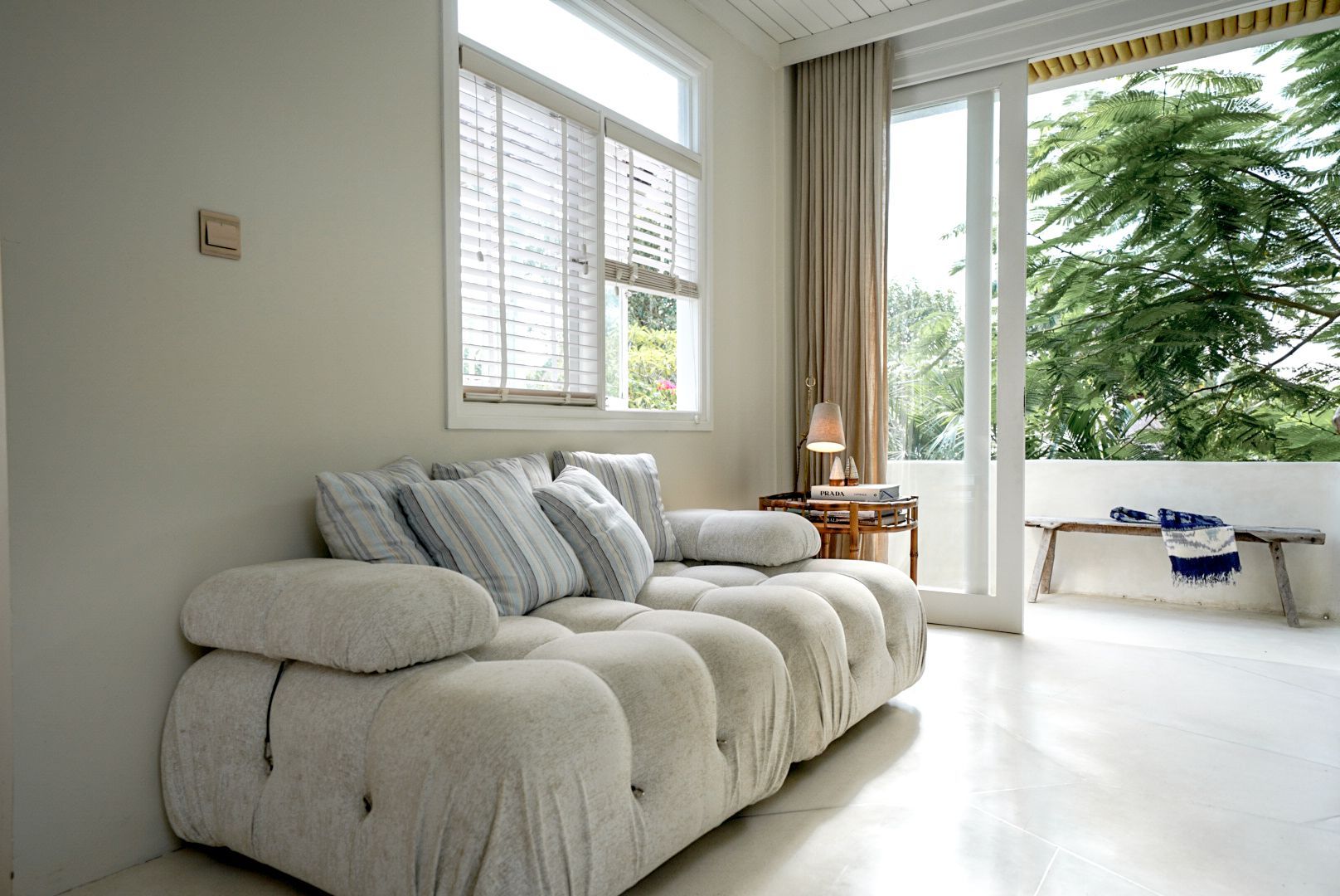 A light-filled room with a white sofa, window with blinds, and open door to a balcony with a bench and greenery.