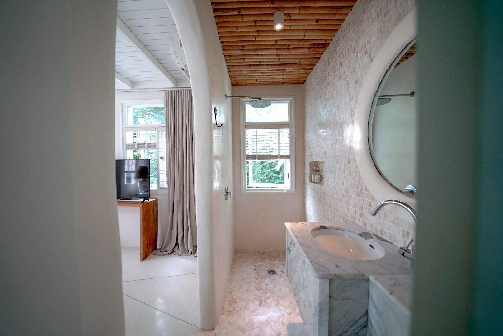 Bathroom interior with a white marble sink and shower, a bamboo ceiling, and a view into a room with a TV and window.