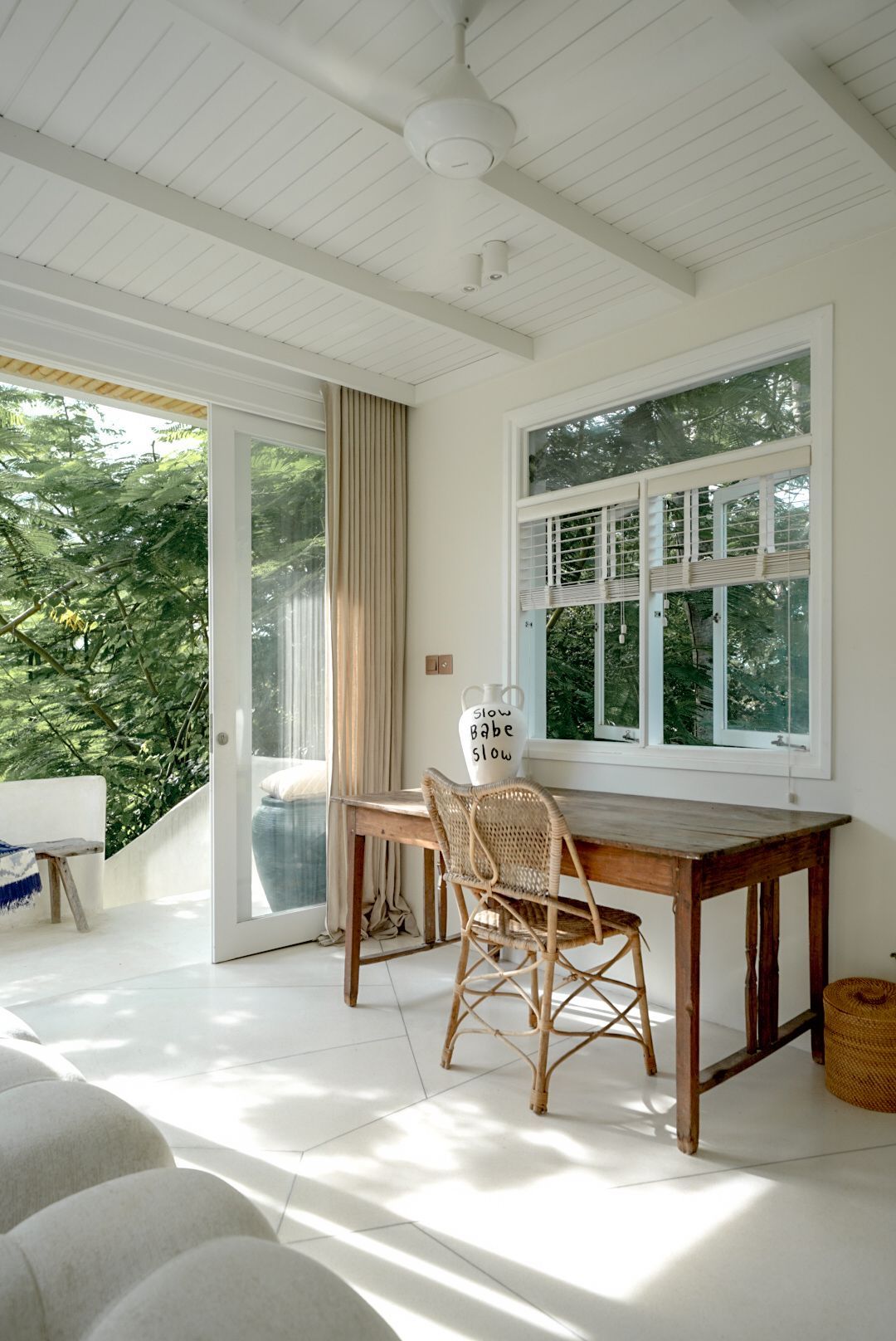 Bright white room with wooden desk and wicker chair by a window, and a doorway opening to lush greenery.