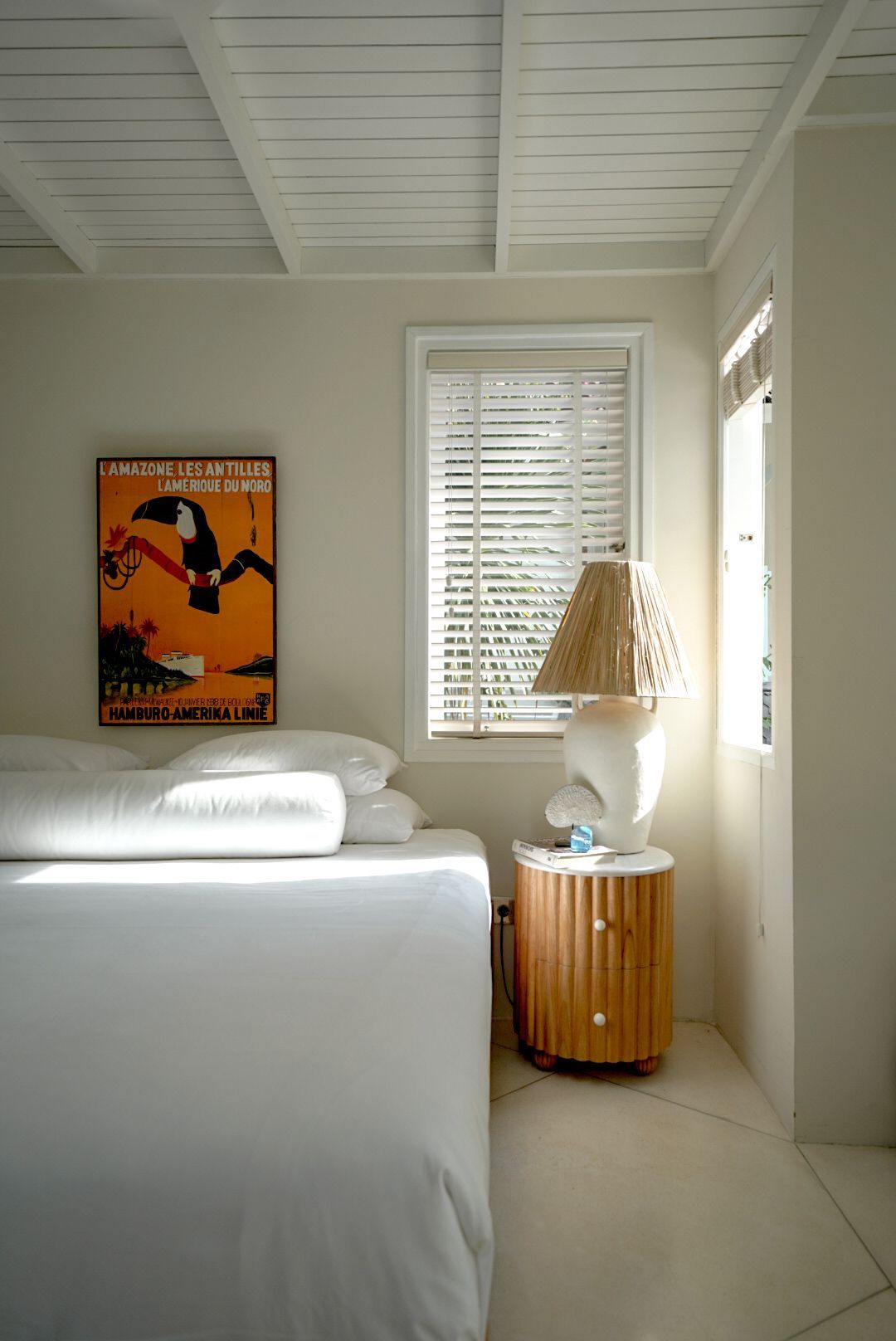 Bedroom with white bedding, a vintage poster, and a bedside table with a lamp. Bright sunlight streams through the window.