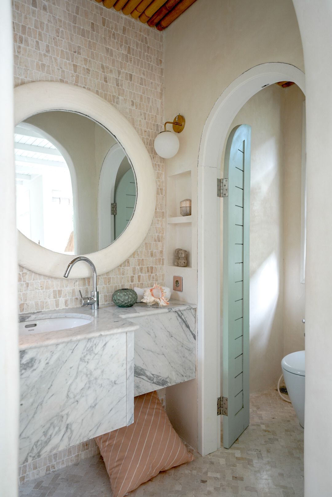 A bright, airy bathroom with a marble sink, large round mirror, and arched doorway leading to a toilet area.