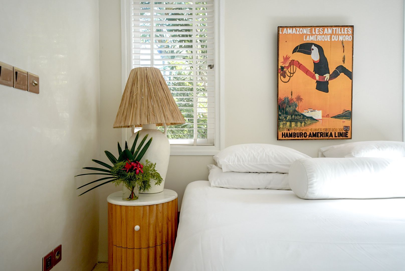 Bedroom with a white bed, a round bedside table, and a framed toucan poster. A window is behind the table.