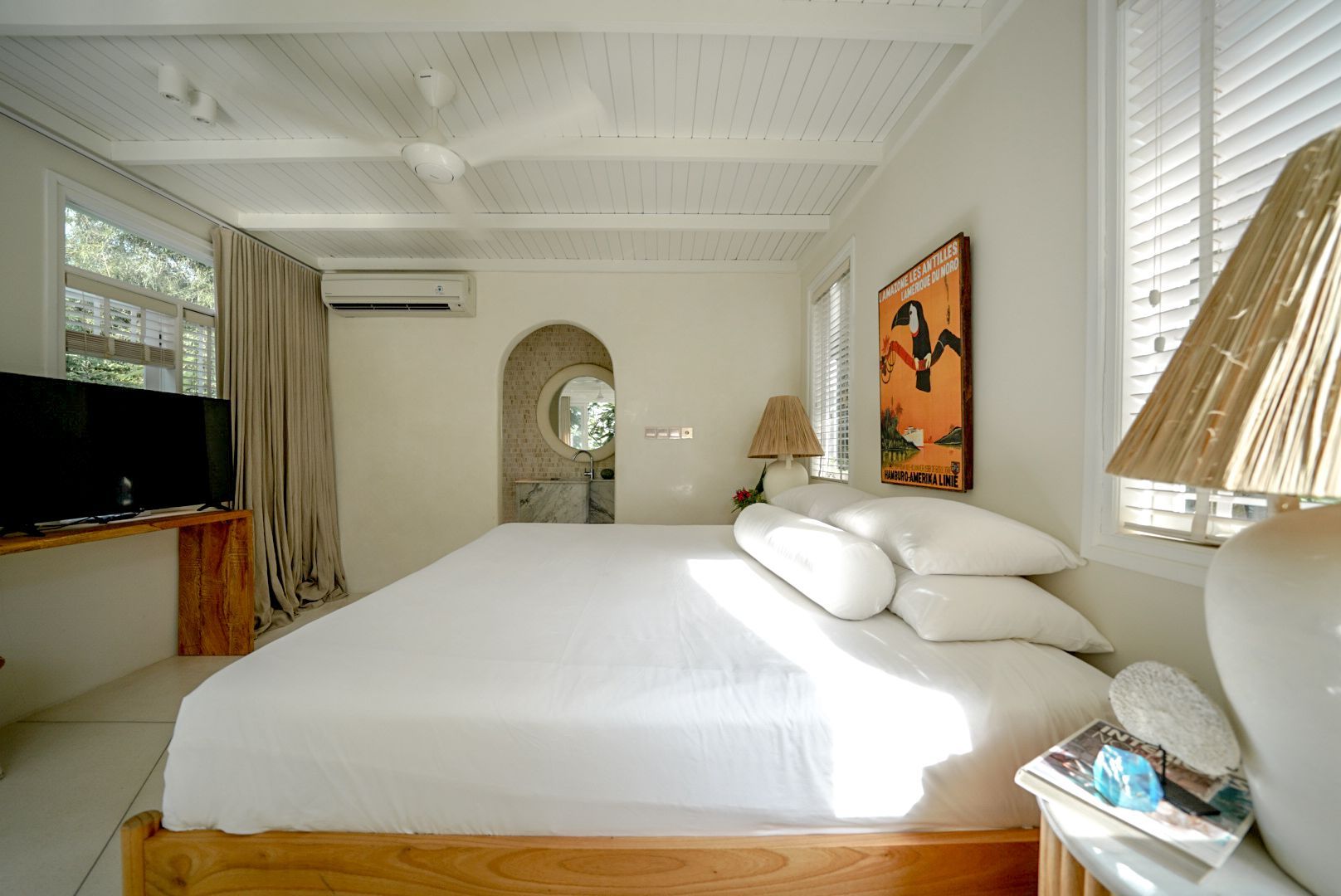 Bright bedroom with white bedding, wooden accents, and a doorway leading to a bathroom. Sunlight streams through windows.