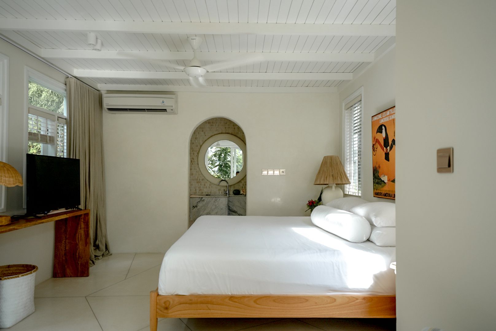 Bedroom with white walls and ceiling, wooden bed, and an arched doorway leading to a bathroom. Sunlight streams in.