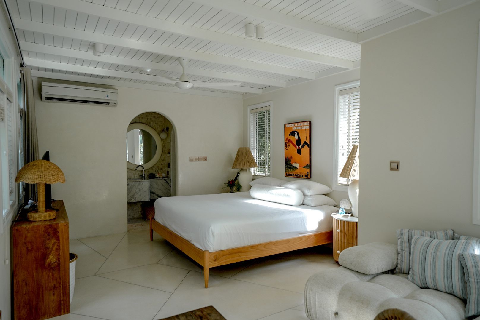 Bright bedroom with white walls, wood ceiling, and bed. A bathroom is visible through an arched doorway.