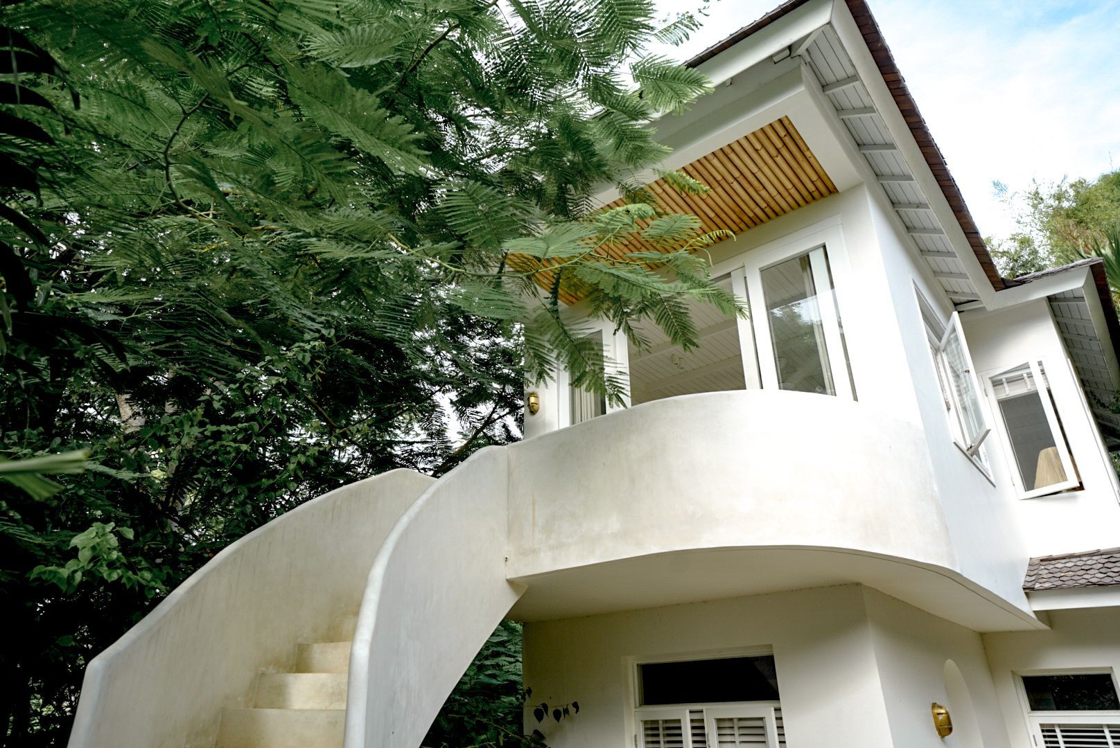 White two-story house with a curved staircase. Tall green trees surround the building, partially obscuring it.