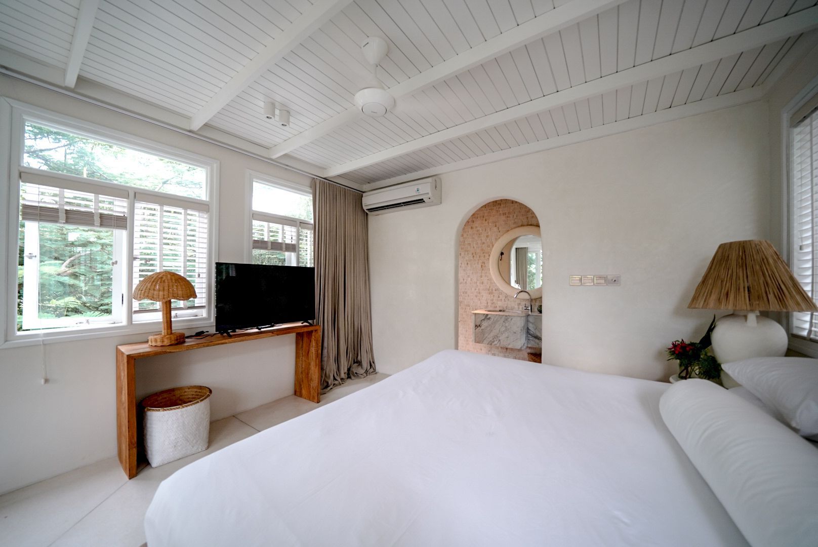 Bright bedroom with white walls, windows, and bed. A wooden console holds a TV and lamp. An archway leads to a bathroom.