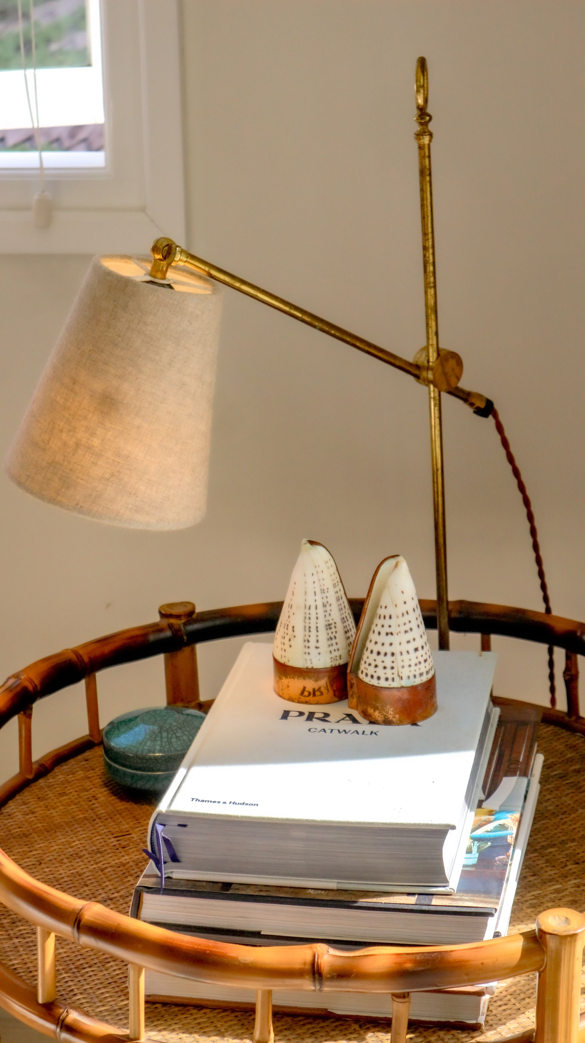 A brass lamp illuminates books and decorative objects on a rattan side table next to a window.