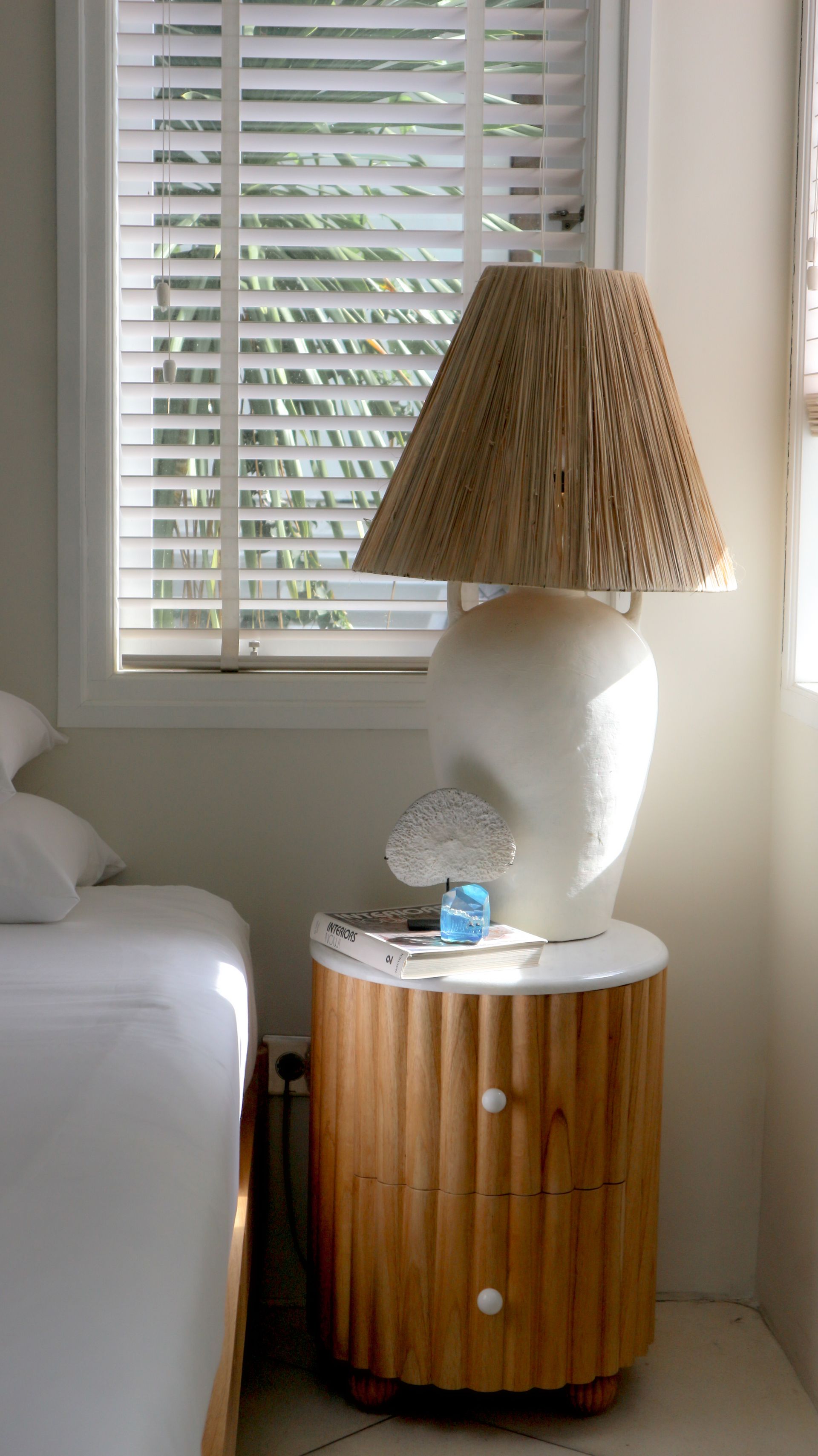 Nightstand with lamp beside a bed near a window. The lamp has a tan lampshade, white base, and sits on a wooden nightstand.