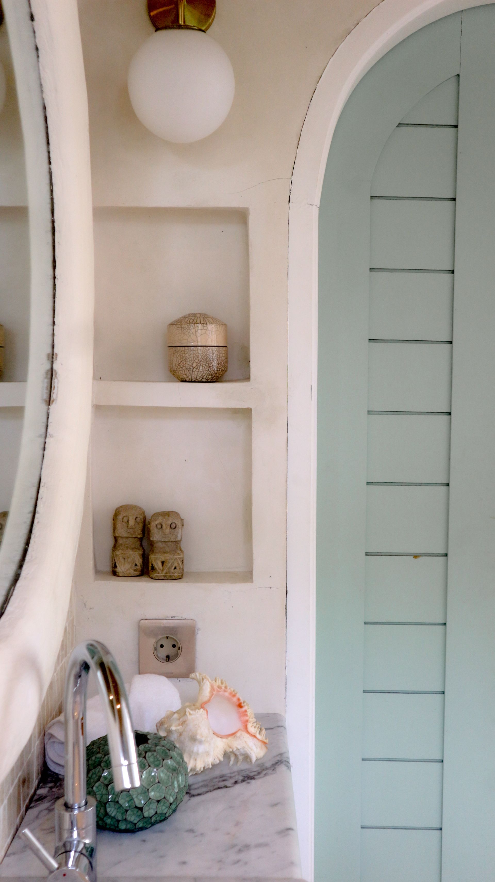 Bathroom vanity with built-in shelves and a light blue arched door. White walls, marble countertop, and gold accents.