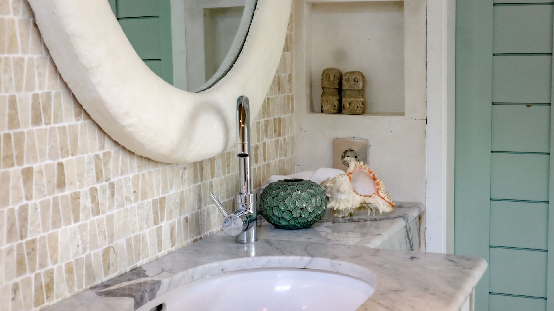 Bathroom with oval mirror, mosaic tile wall, marble countertop, and seashell decor. Features a round sink and modern faucet.