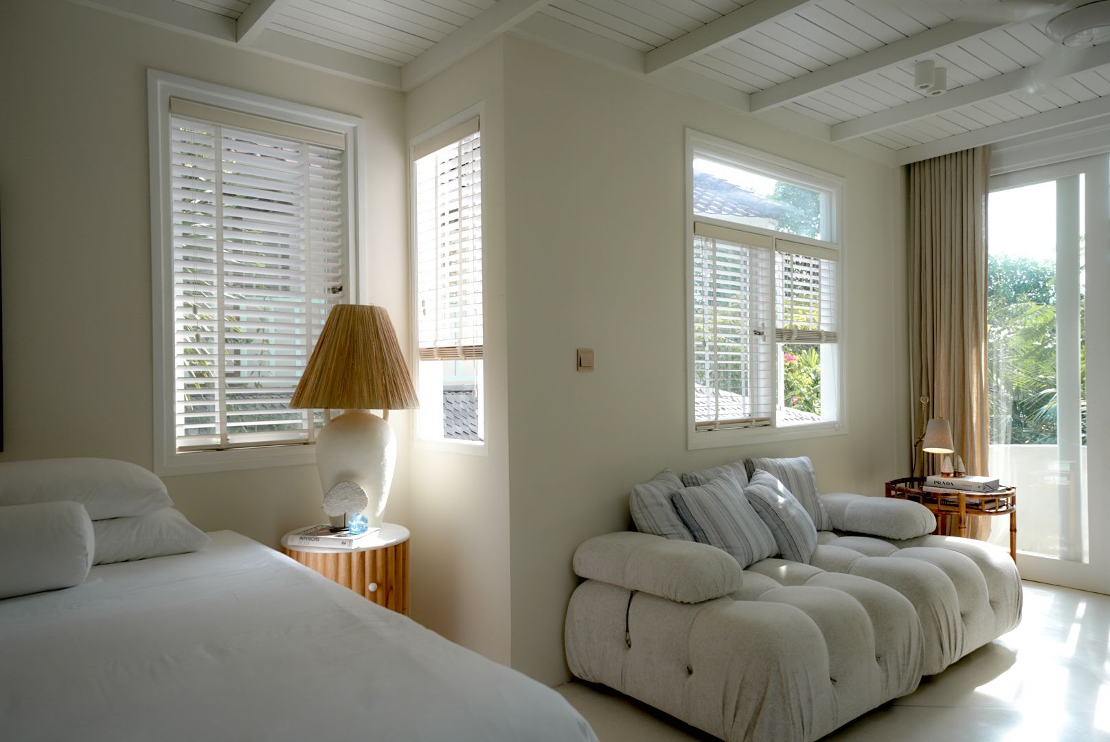 Bedroom with white walls, windows with blinds, bed, and a tufted white sofa. Sunlight streams in, illuminating the room.
