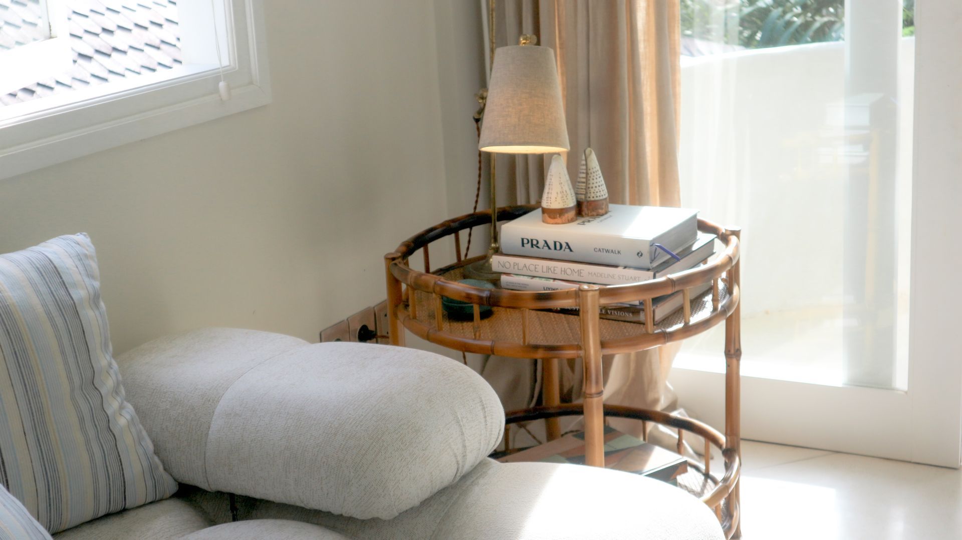 A cozy living room setting with a round bamboo side table holding books and a lamp next to a white couch.