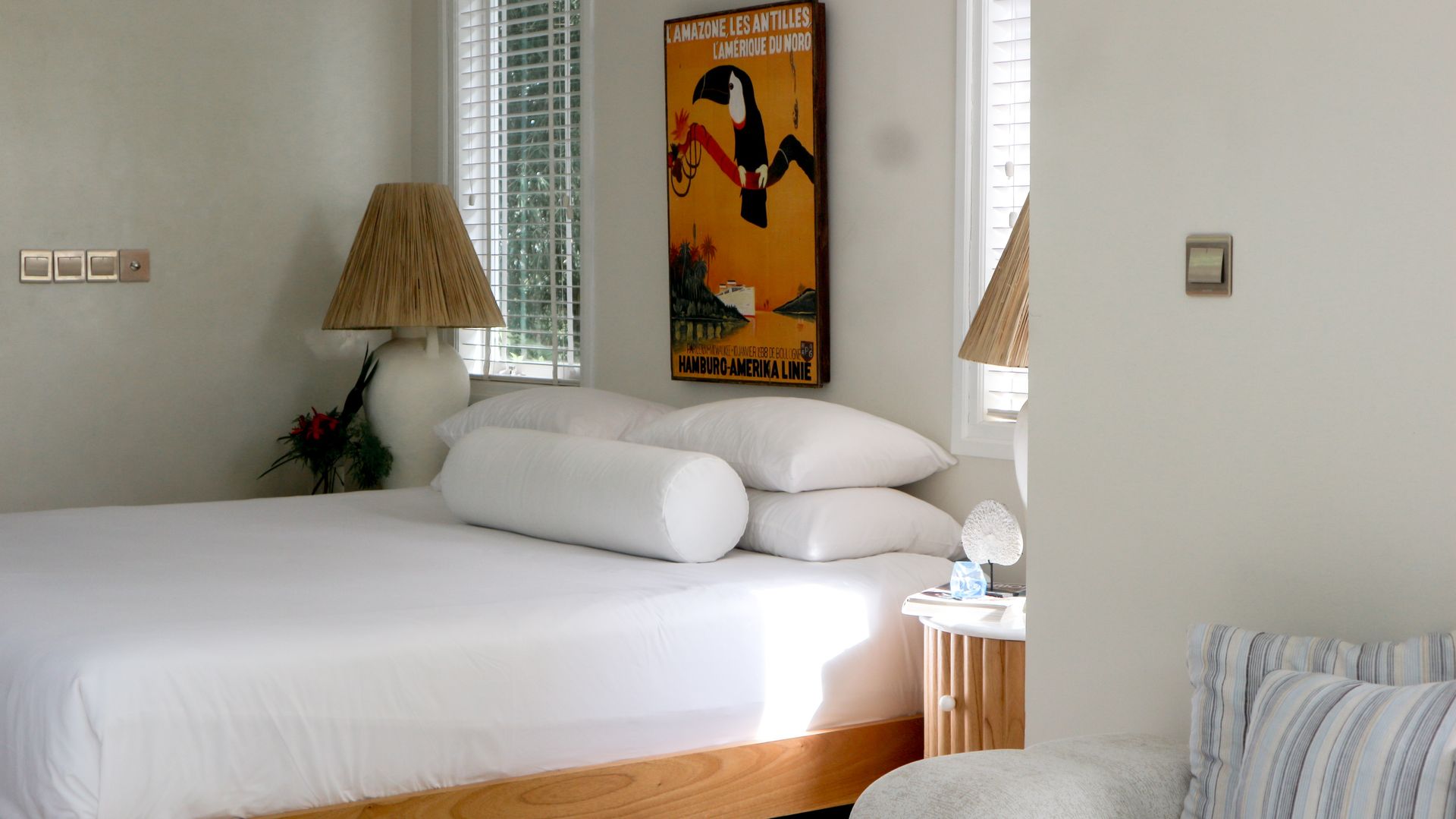 A well-lit bedroom with a bed, lamps, artwork, and a small side table. The bed has white bedding.