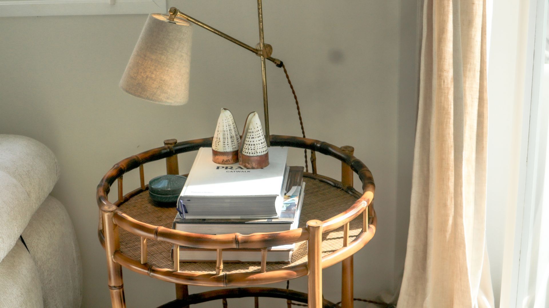 A round, bamboo side table with books, decorative objects, and a lamp in a bright room.