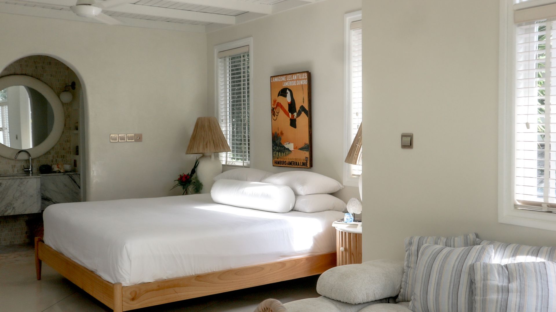 A bright, neutral bedroom with a wooden bed, artwork, and a small seating area. Sunlight streams in through the windows.