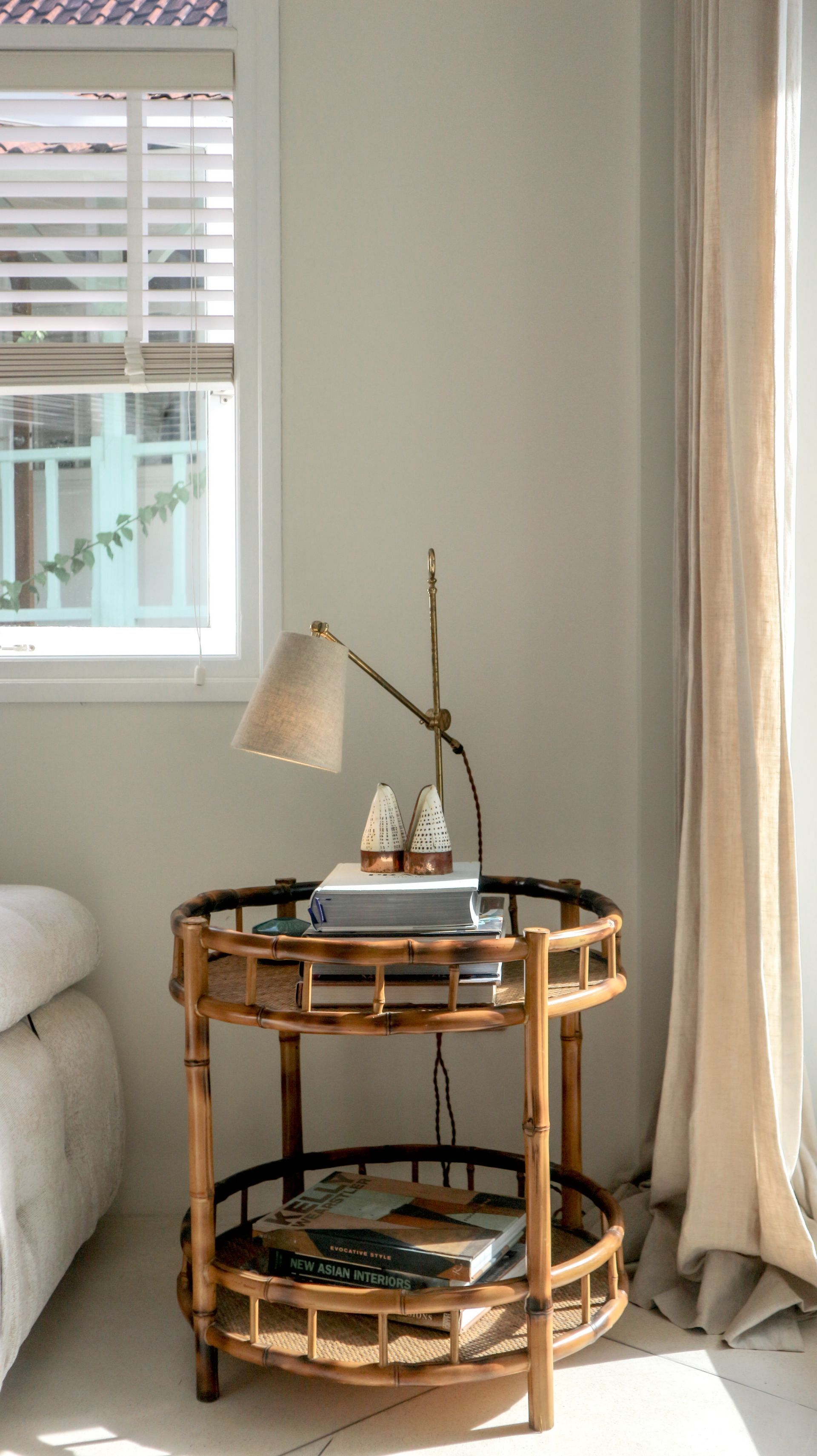 A round, bamboo side table with books and a lamp sits by a window with a view of a light blue building.