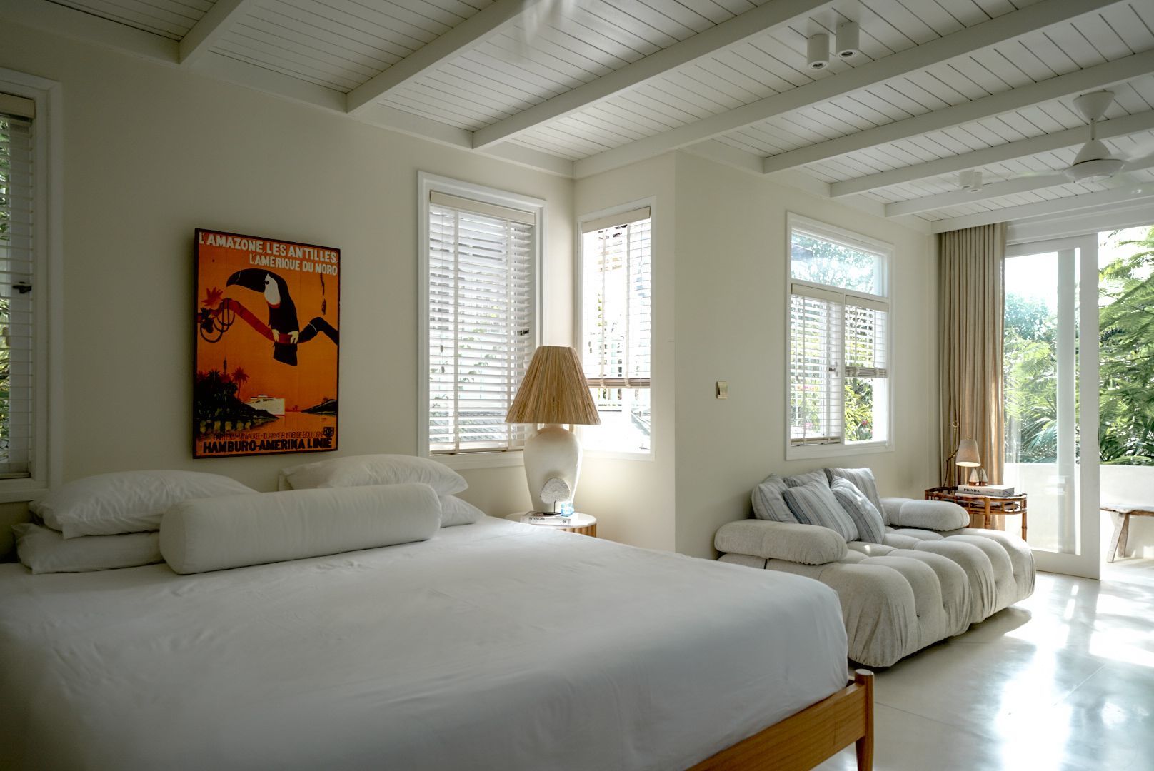 Bright bedroom with a large bed, sofa, and windows. An orange poster hangs above the bed, and sunlight streams in.