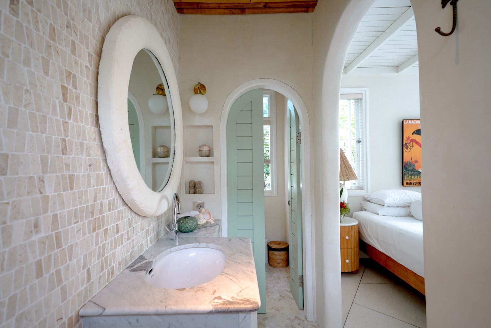 Bathroom with mosaic tile wall, oval mirror, marble countertop, and arched doorway leading to a bedroom.
