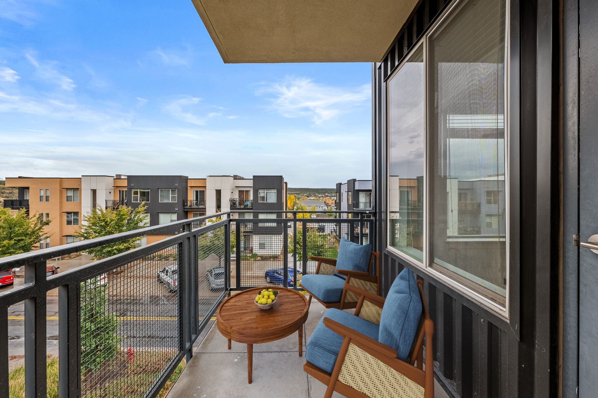 Balcony seating with two blue-cushioned chairs and a round wooden table near sliding glass doors.
