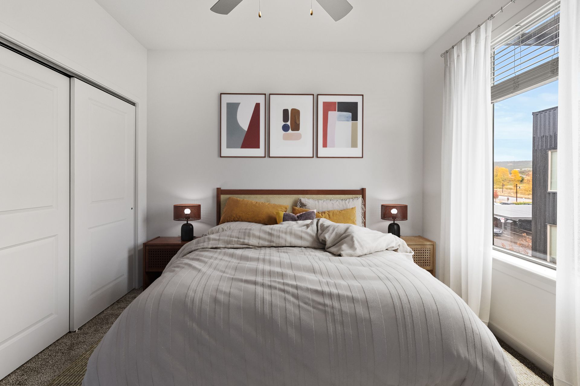 Bedroom with a queen bed, two nightstands, abstract art above the headboard, and a large window with white curtains.