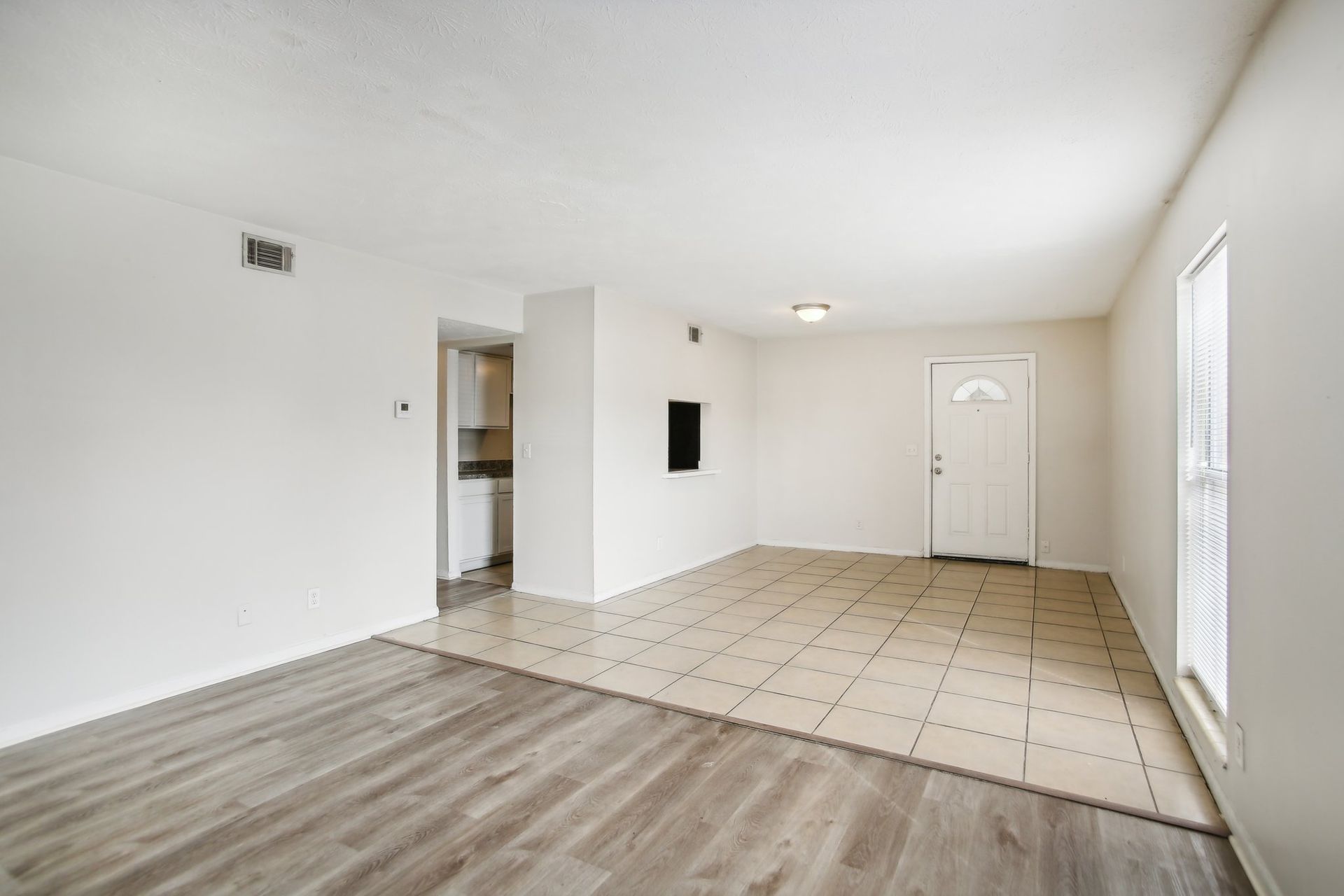Empty, bright living room with tile and wood flooring, white walls, and a doorway to a kitchen area.