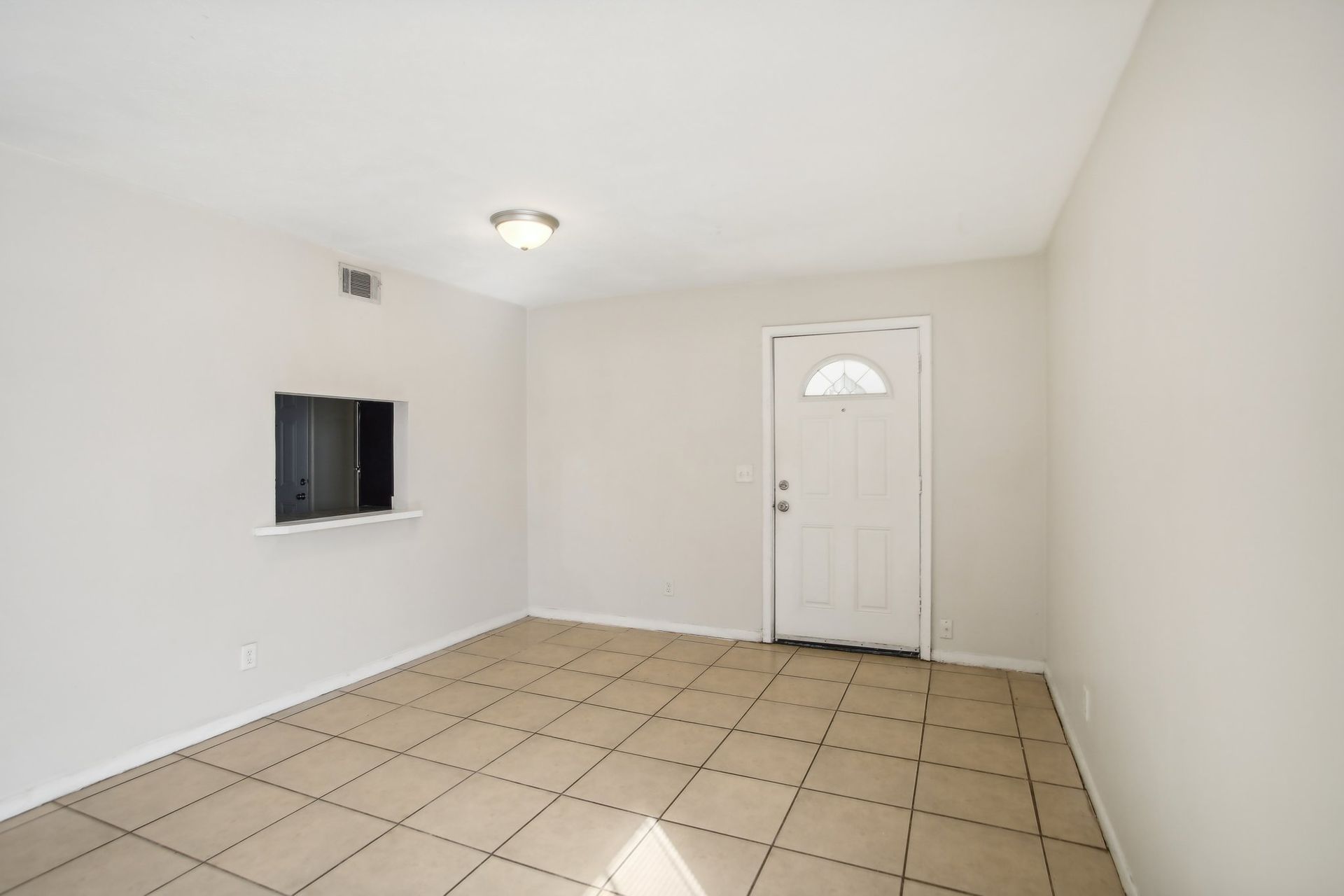 Empty room with tan tiled floor, white walls, small window, white door, and overhead light.