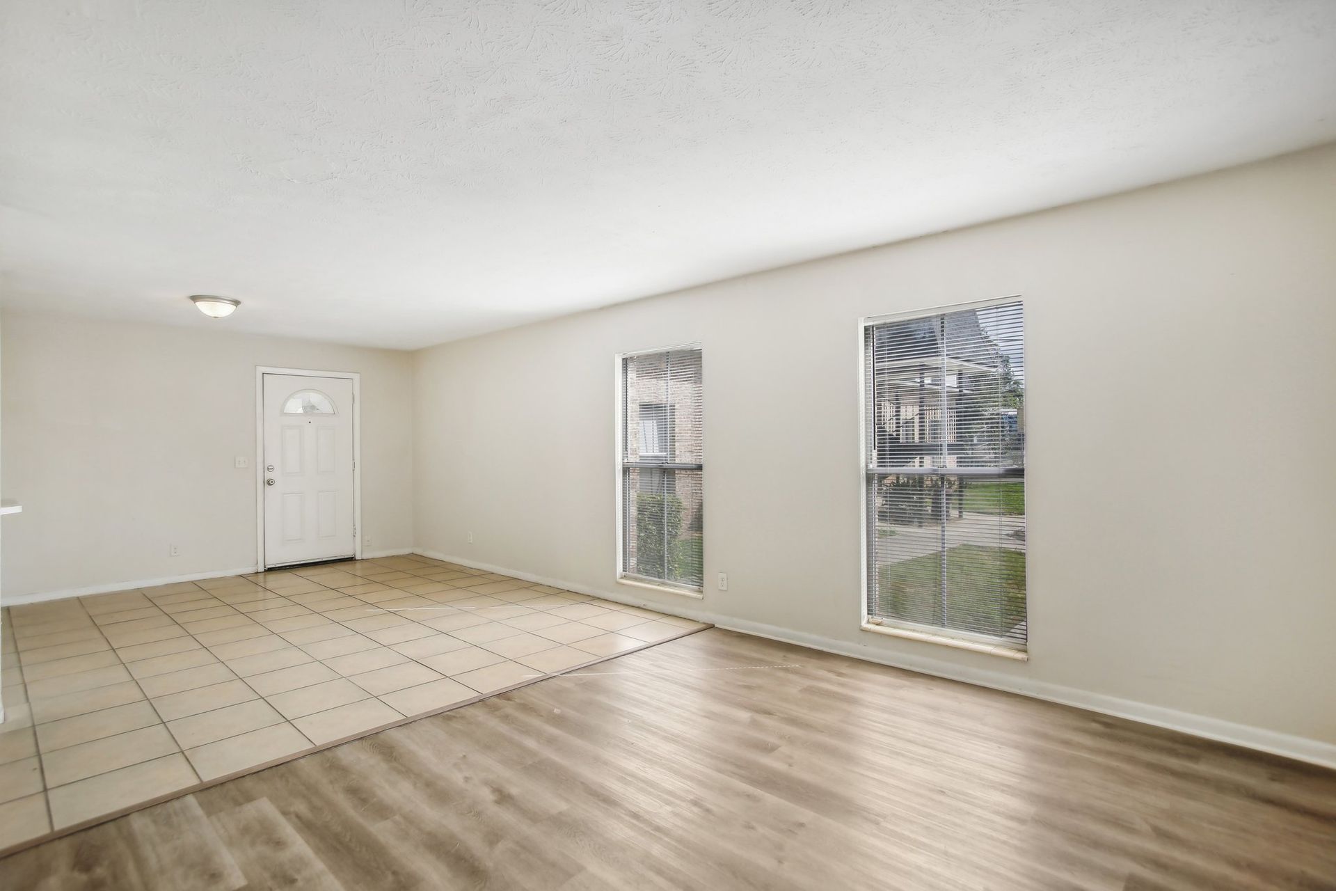 Empty living room with wood-look flooring and tile, white walls, windows, and a doorway.