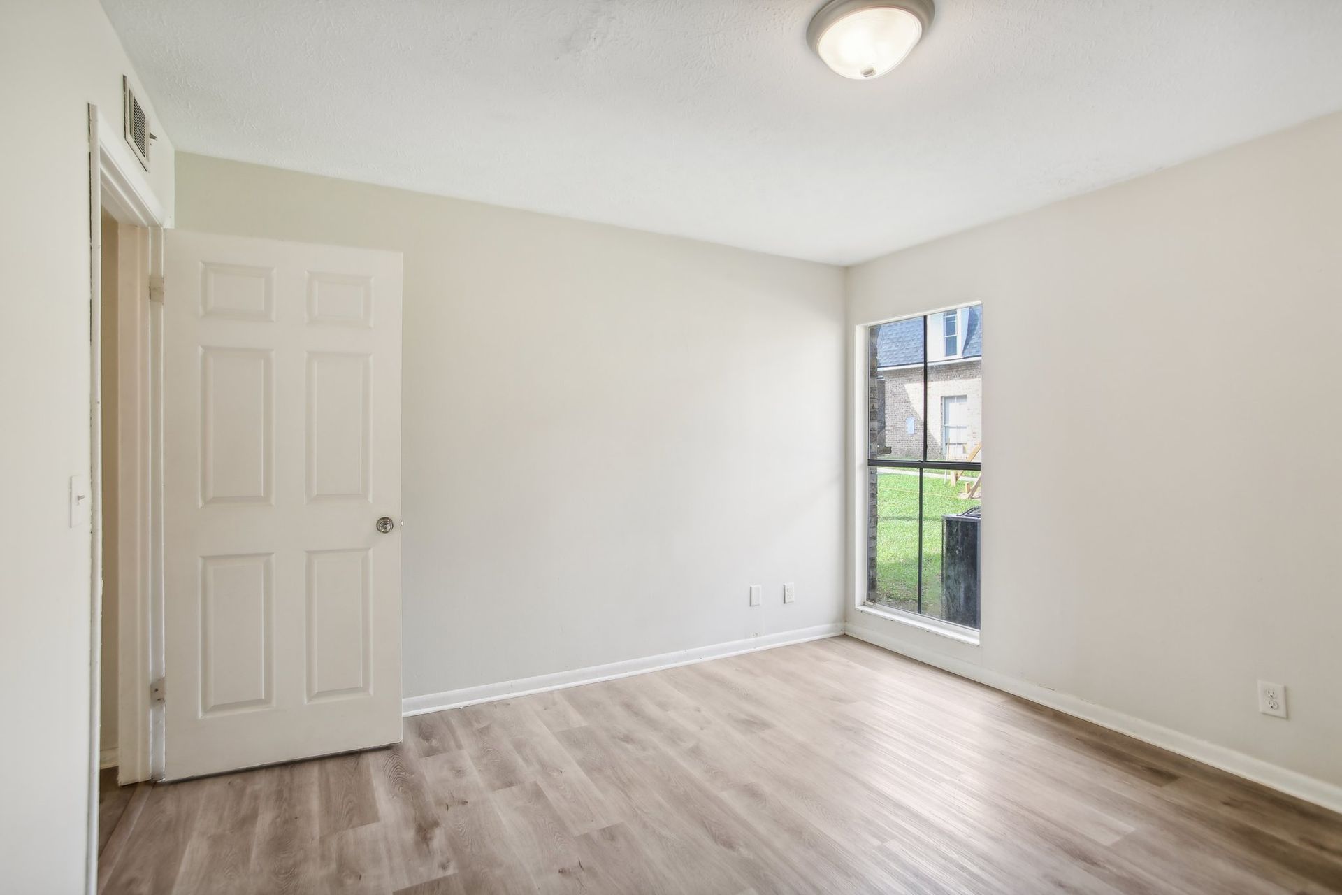 Empty room with gray-white walls, white door, window, light wood floor, and overhead light.