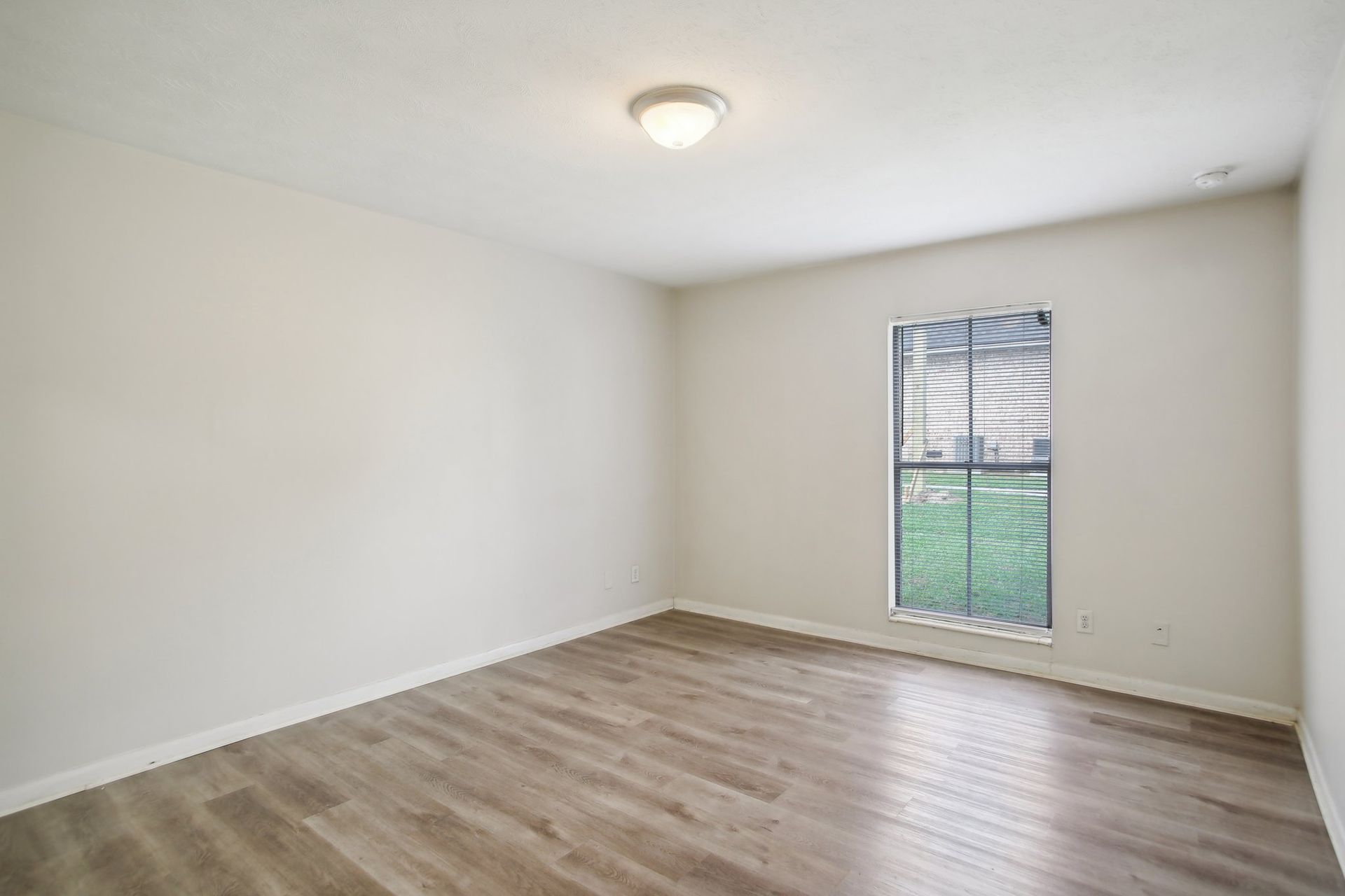 Empty room with wood-look floor, window with blinds, and a ceiling light. White walls.