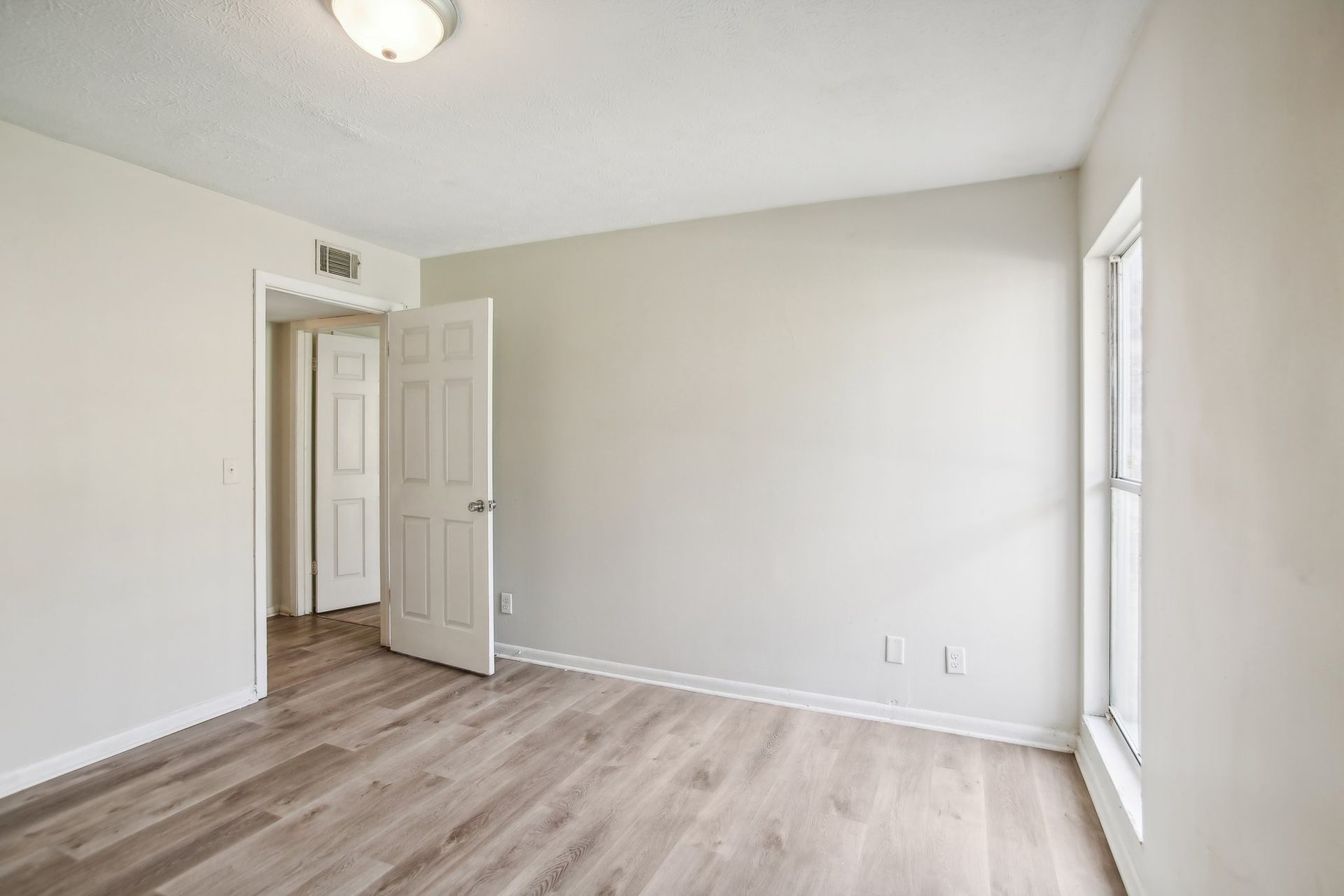 Empty bedroom with pale gray walls, wooden floors, open door to hallway, and window.