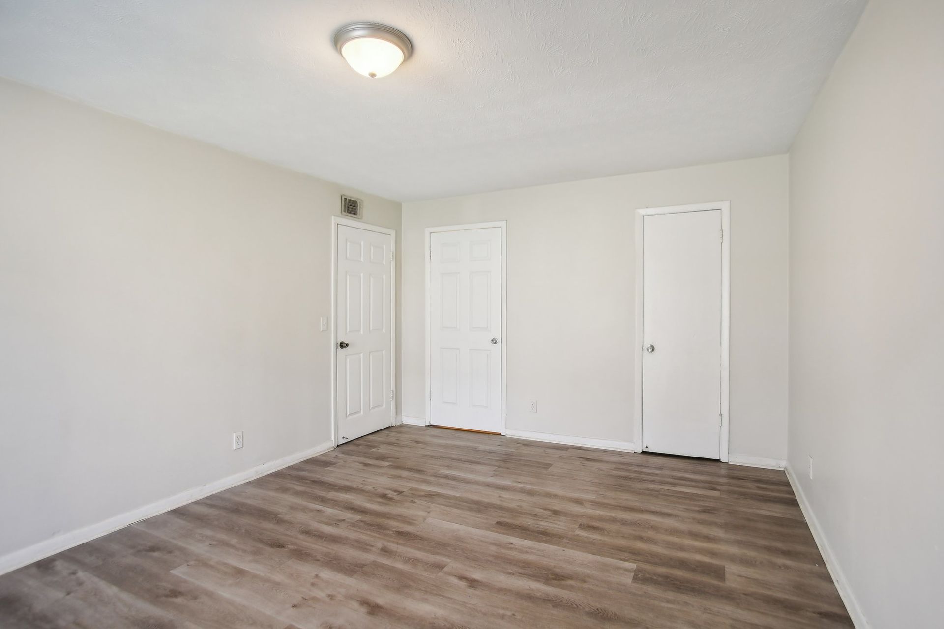 Empty room with light-colored walls, three white doors, and wood-look flooring. A ceiling light illuminates the space.