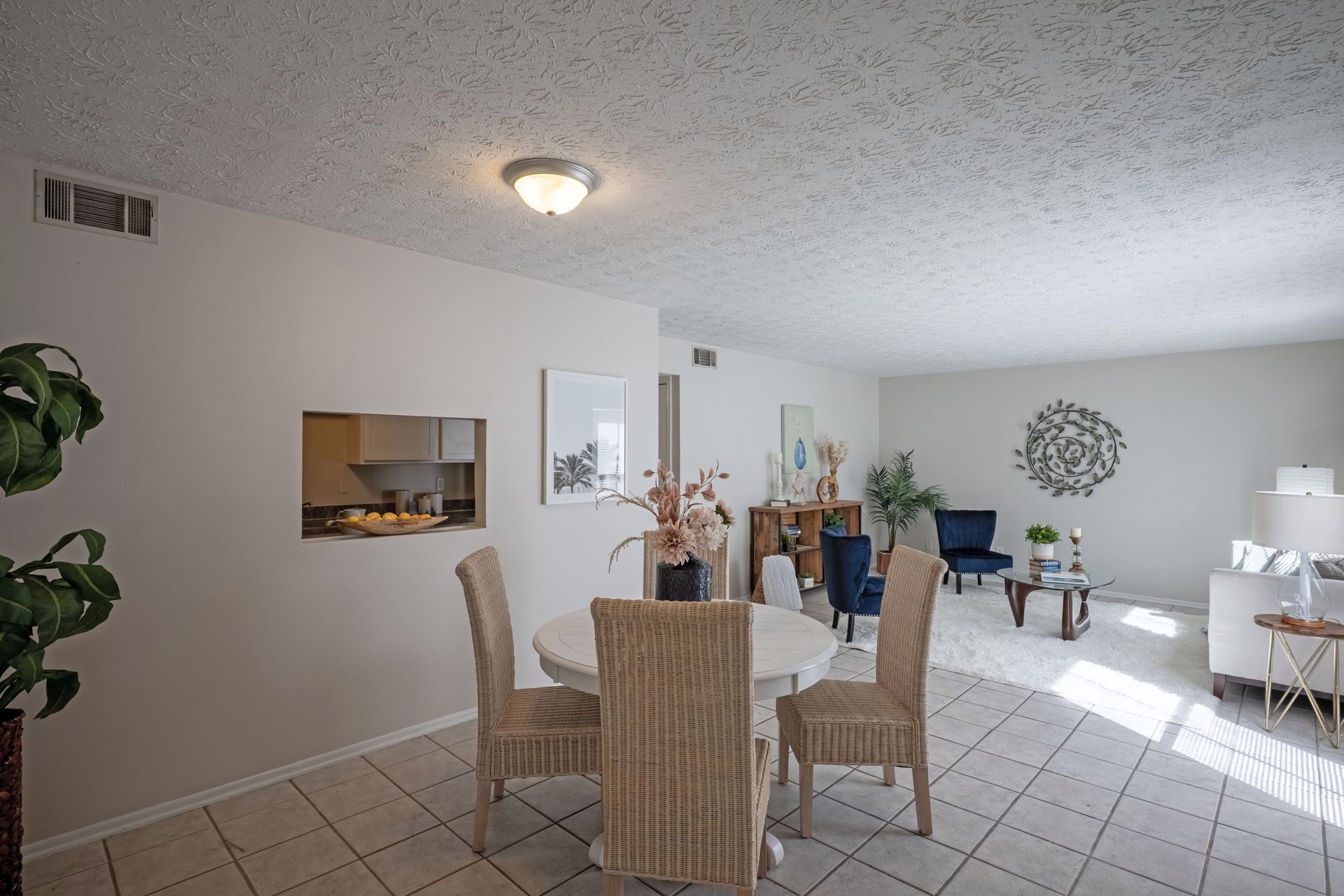 Dining area with a round table, wicker chairs, and a living room in the background. White walls, tile floor.