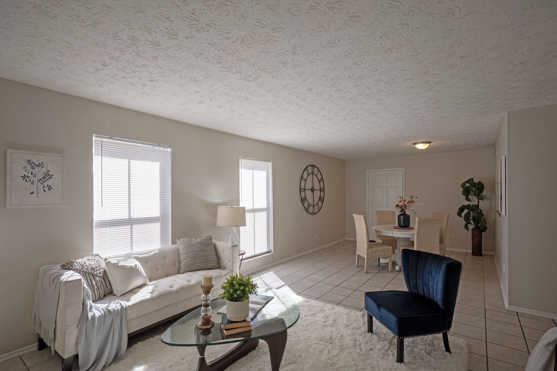 Living room with couch, coffee table, windows, dining table, and blue chair. Bright with white walls and rug.