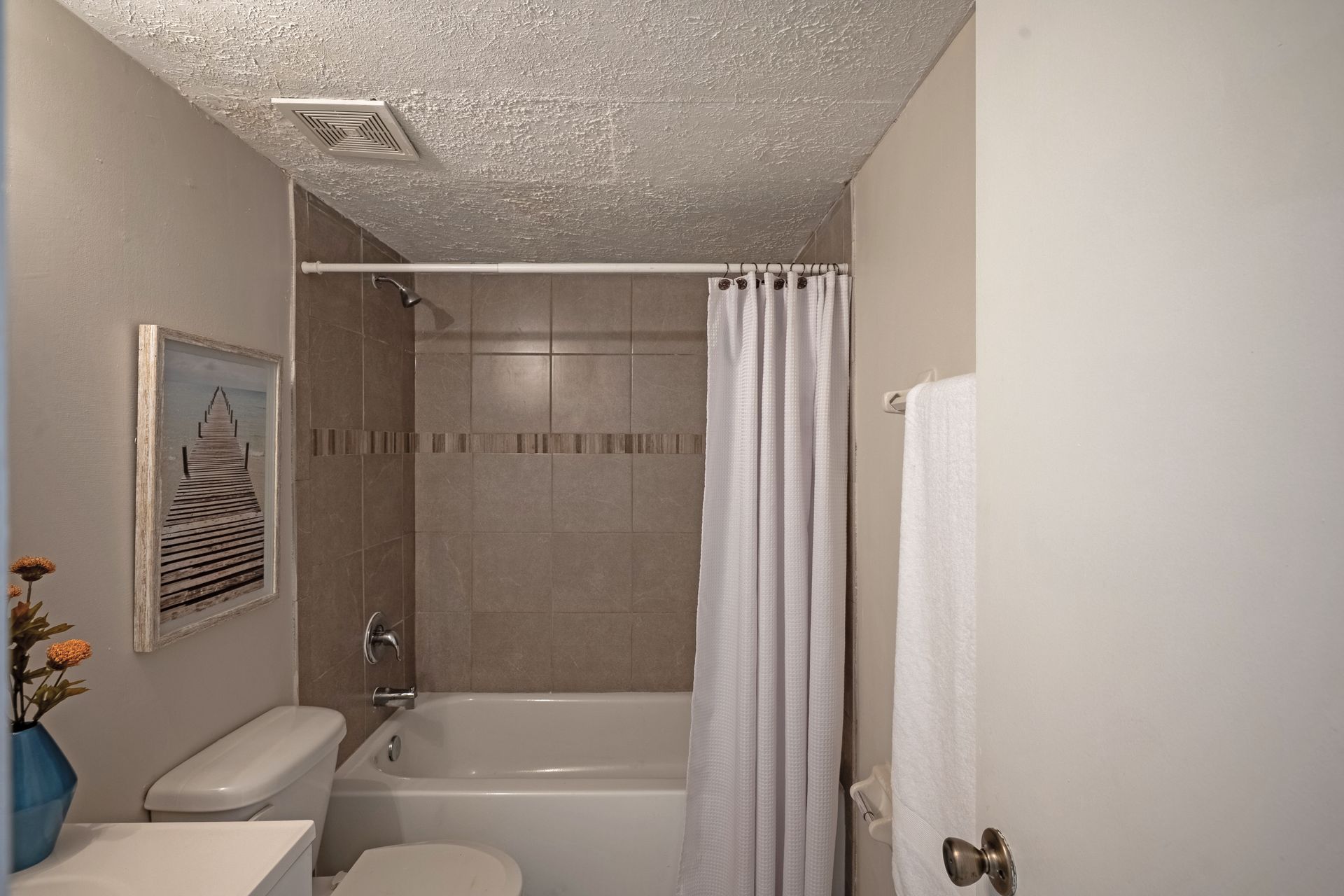 Small bathroom with toilet, bathtub, and shower curtain. Beige walls, textured ceiling, and a framed picture.