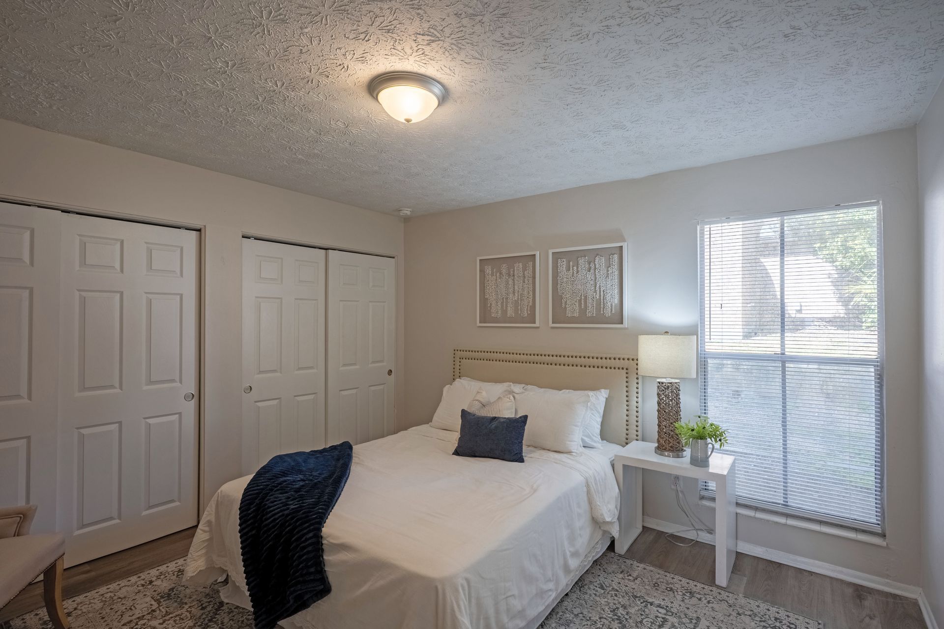 Bedroom with white walls, bed, and closed closet doors.  Small bedside table with lamp.  Window with blinds.