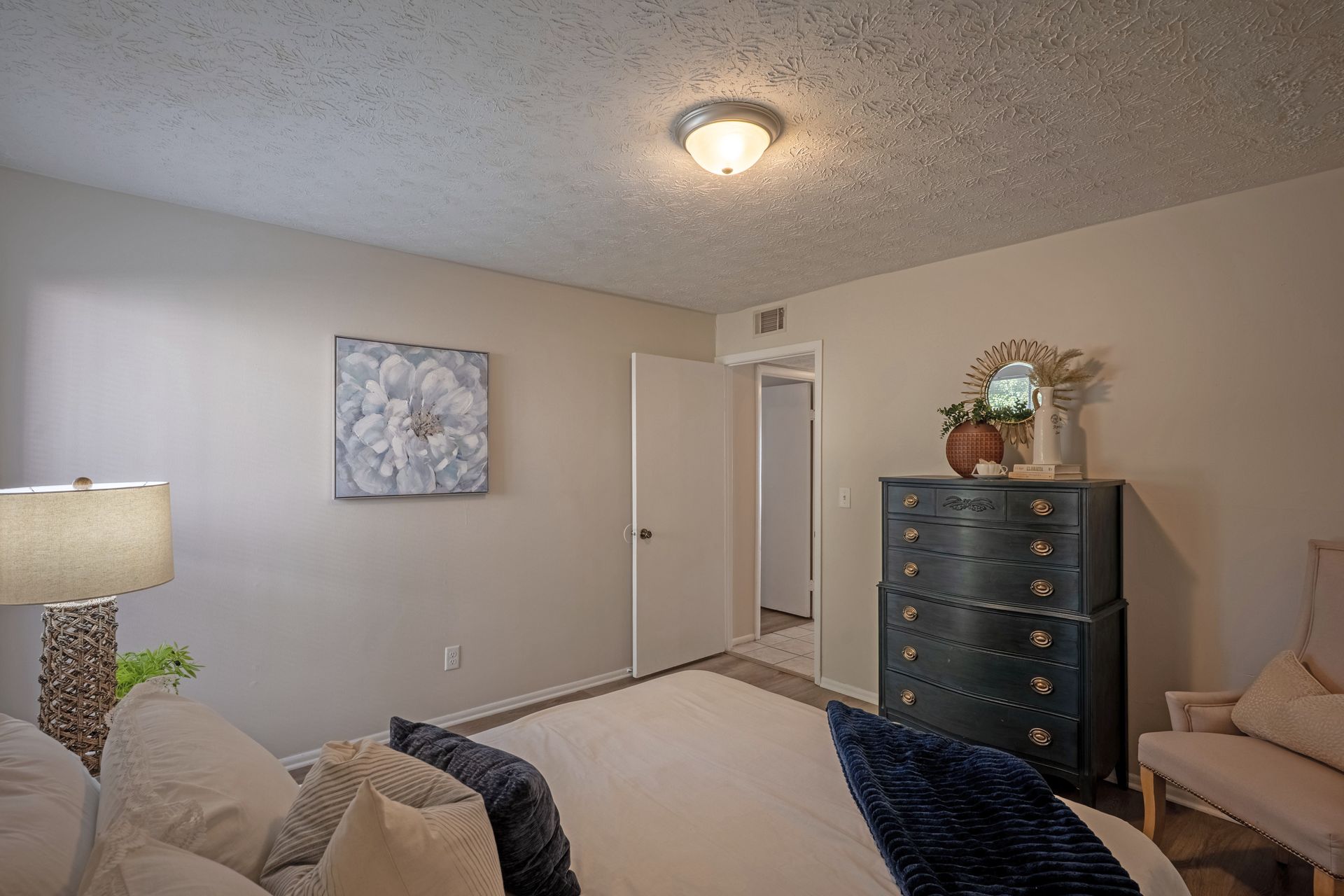 Bedroom with bed, dresser, chair, artwork, and open doorway. Beige walls and textured ceiling.