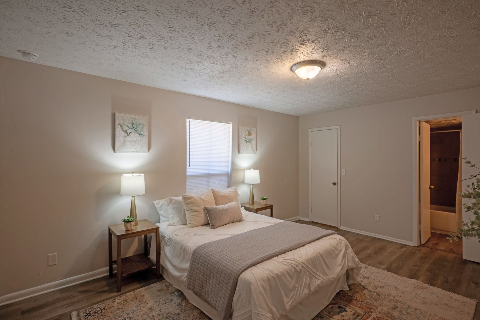 Bedroom with bed, nightstands, lamps, and a window. Light-colored walls and flooring. Door leads to bathroom.