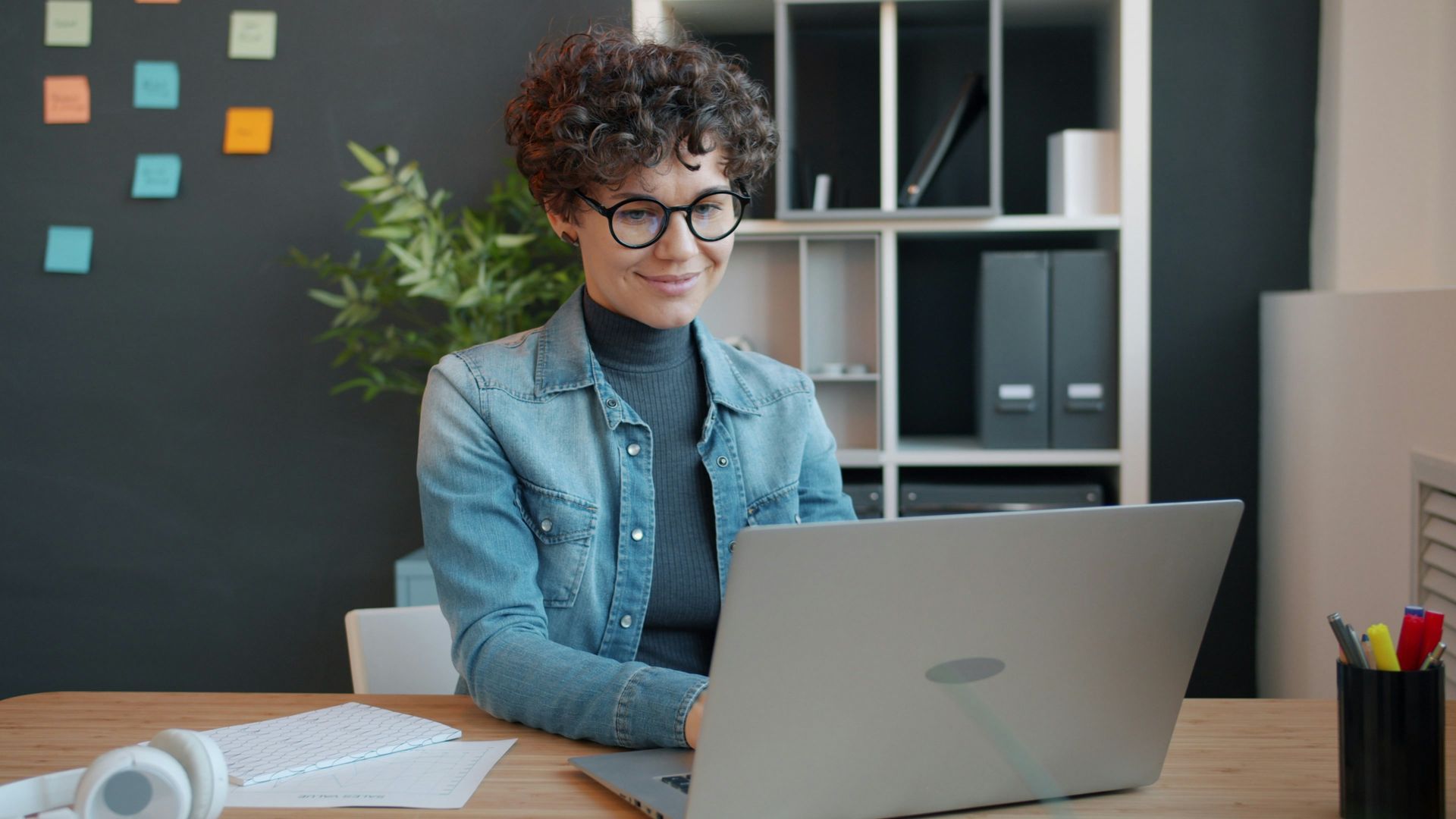 Woman with glasses and curly hair working on a laptop at a desk, smiling.