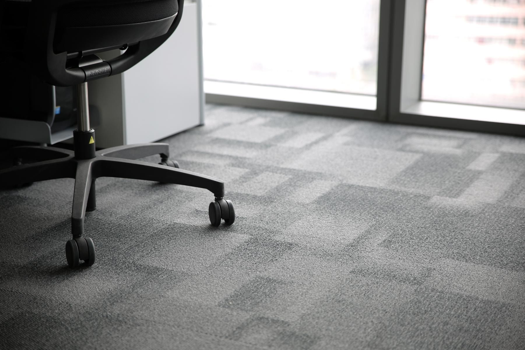A black office chair on wheels sits on a gray patterned carpet next to a bright window in an office.