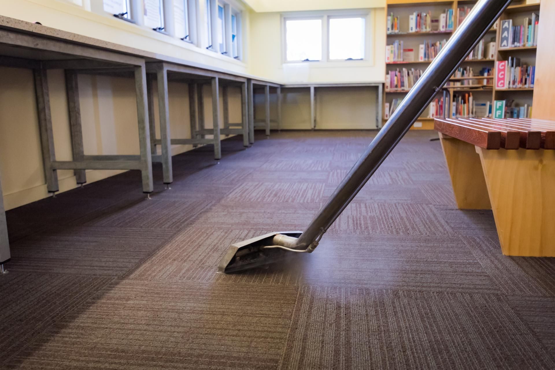 A carpet cleaning wand cleans a square-patterned brown carpet in a room with bookshelves and tables.