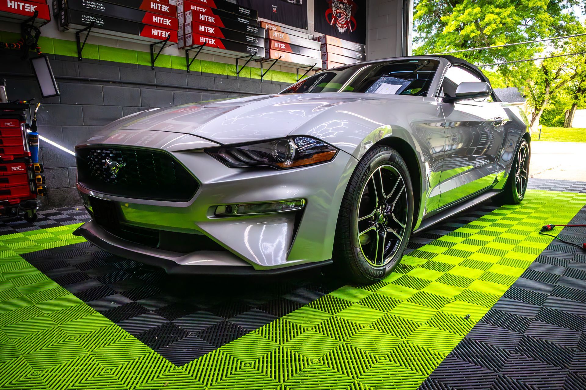 Silver Ford Mustang convertible parked on a green and black tiled floor, under bright lights.